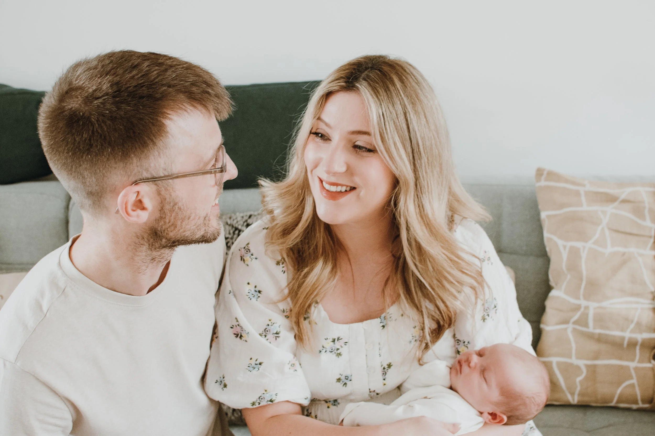 A family consisting of a man with glasses, a woman with wavy blonde hair, and a newborn baby, sitting on a couch with decorative pillows in the background.