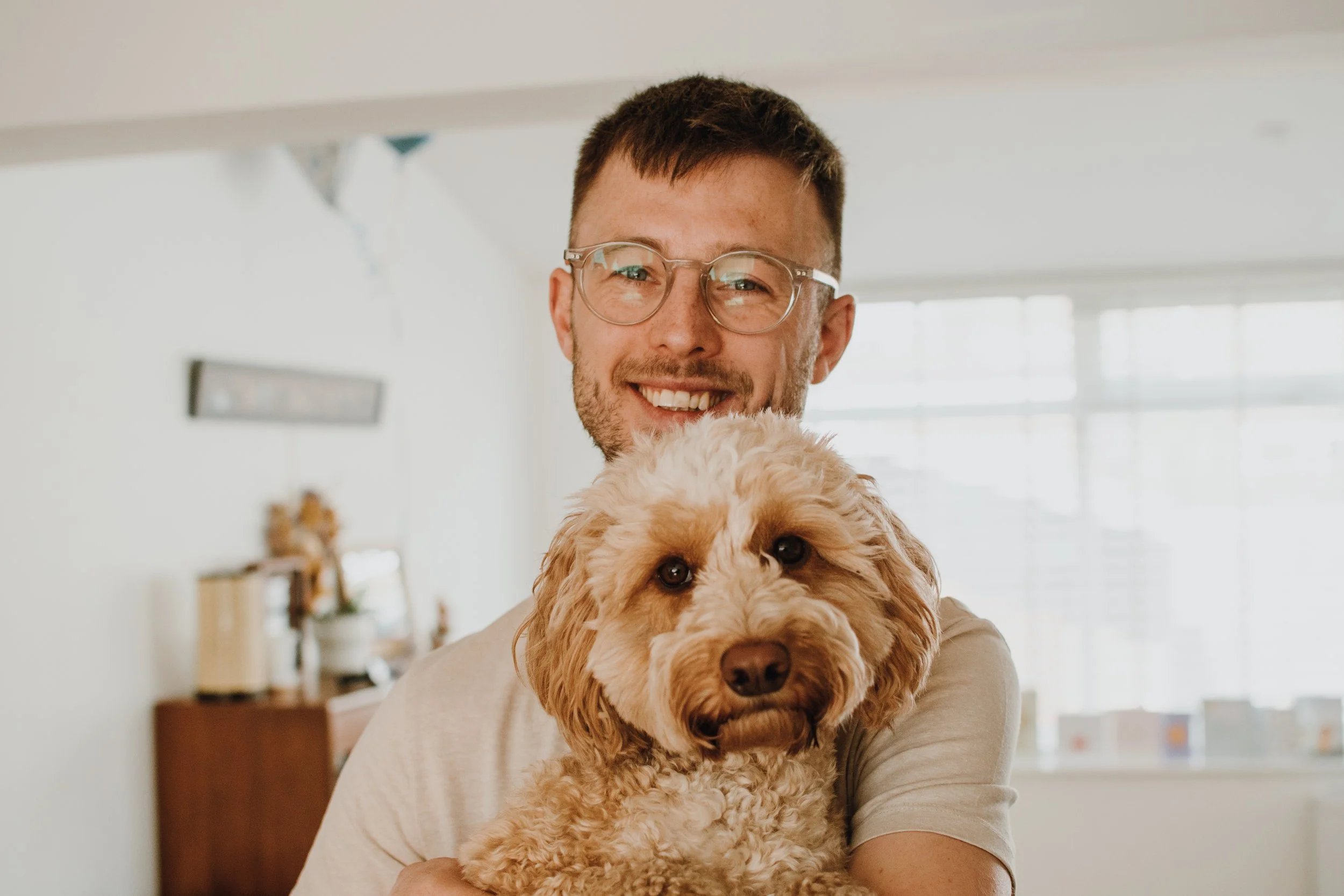 A man with glasses holding a brown, curly-haired dog inside a bright, home interior.