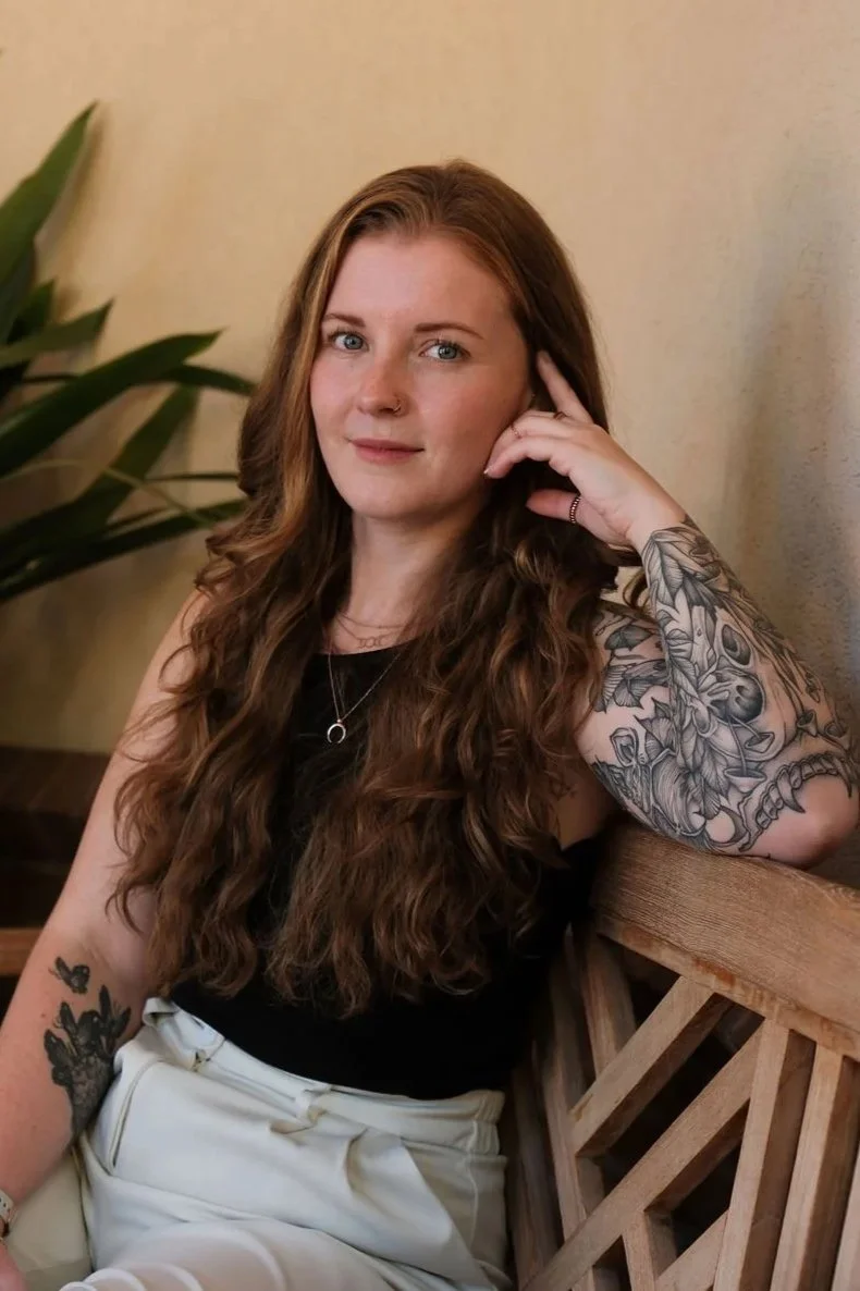 Portrait of a smiling woman with long wavy reddish-brown hair and floral tattoos sitting in a wooden chair beside a houseplant.