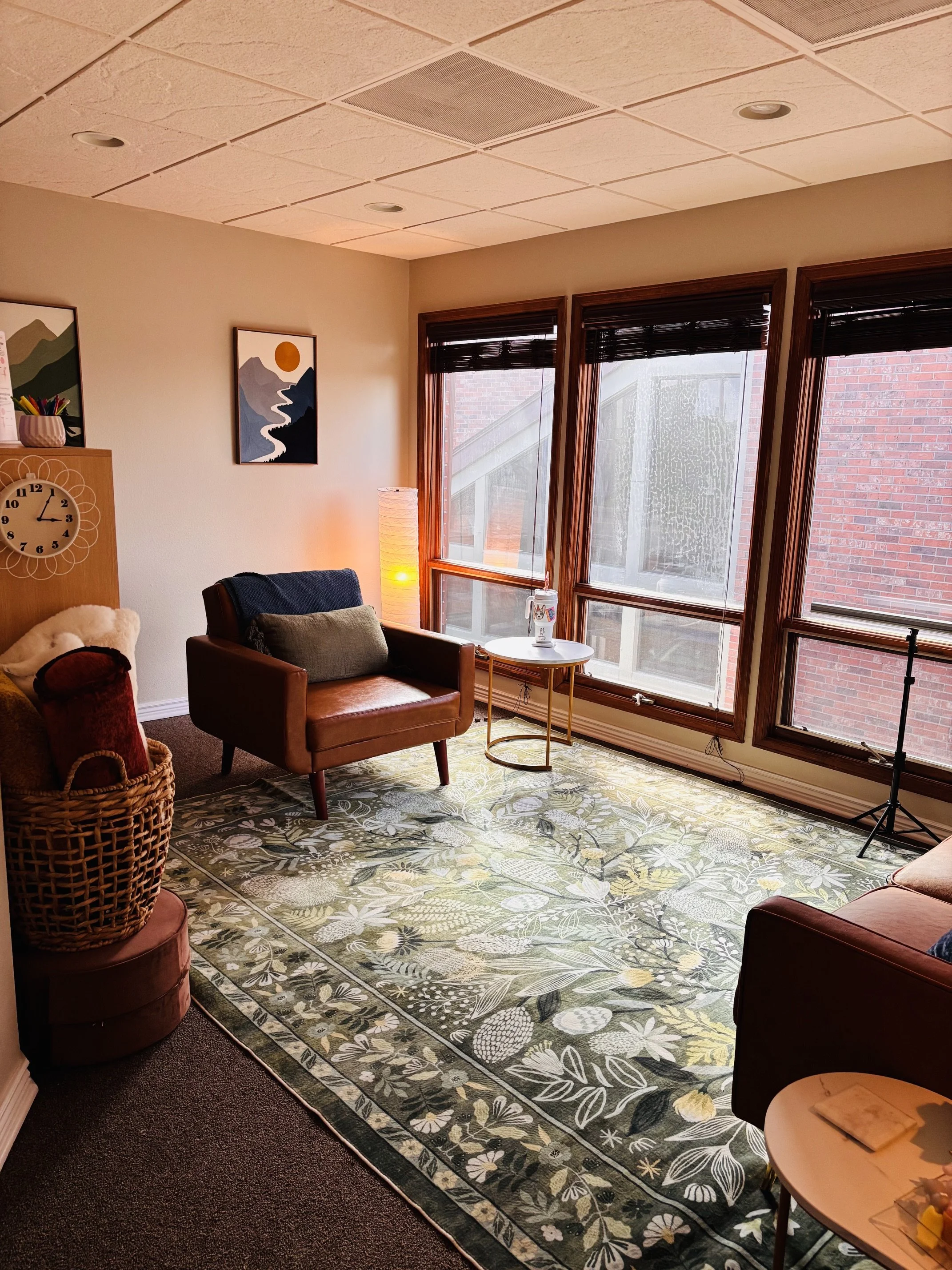Cozy office with a leather armchair, floral area rug, floor lamp, framed artwork, large windows with blinds, and a woven basket.