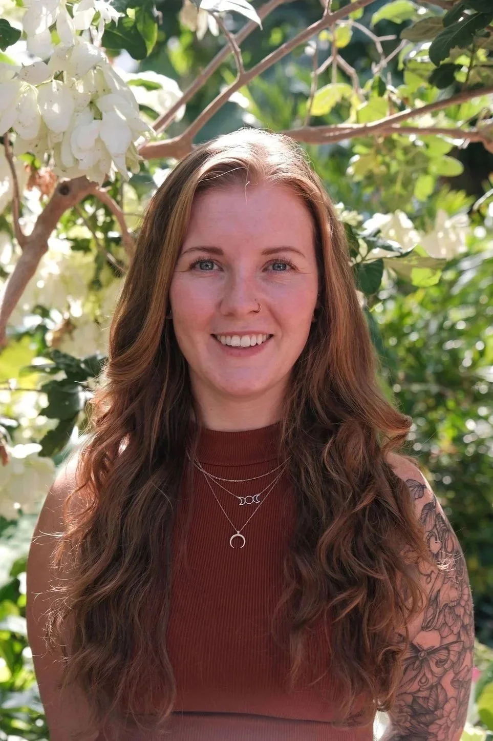 Smiling woman with long reddish-brown hair and a floral tattoo standing outdoors beneath white flowers