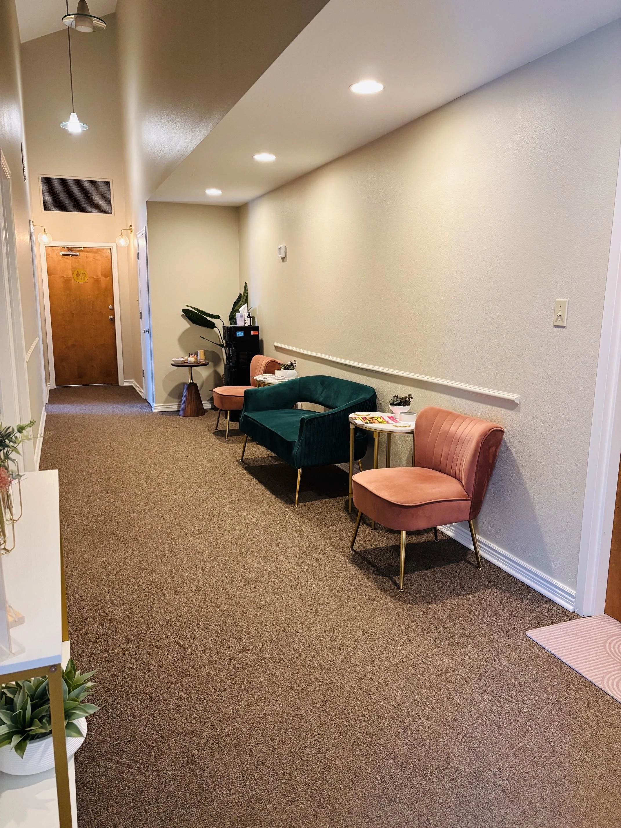 Cozy waiting room with pink chairs, a green loveseat, side tables, plants, coffee station, and restroom door