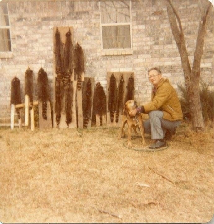 A man sitting on a tree root with a redbone coonhound, near a brick wall with hanging raccoon pelts and a window, in an outdoor yard.
