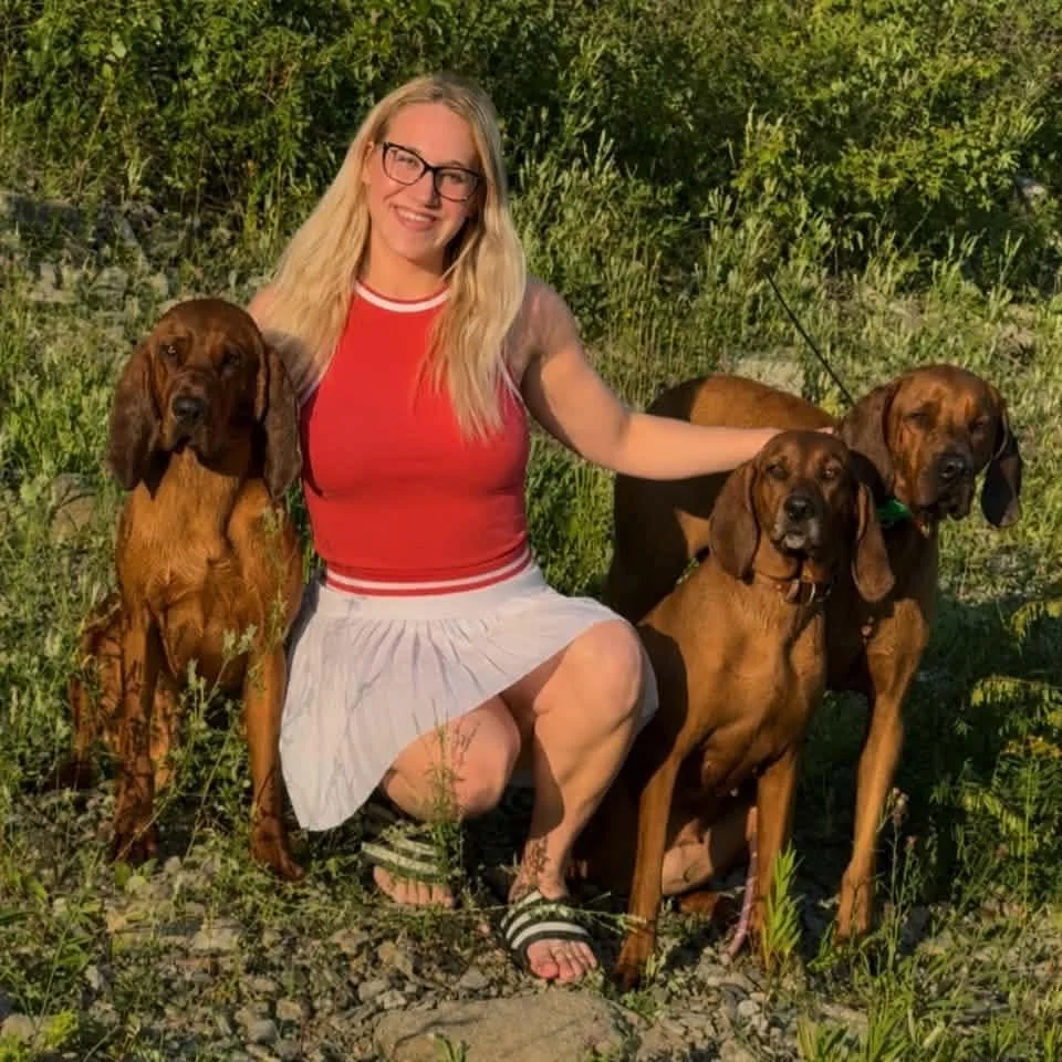 A woman with blonde hair and glasses kneeling outdoors on grassy terrain, surrounded by three Redbone Coonhounds of different genders and ages.