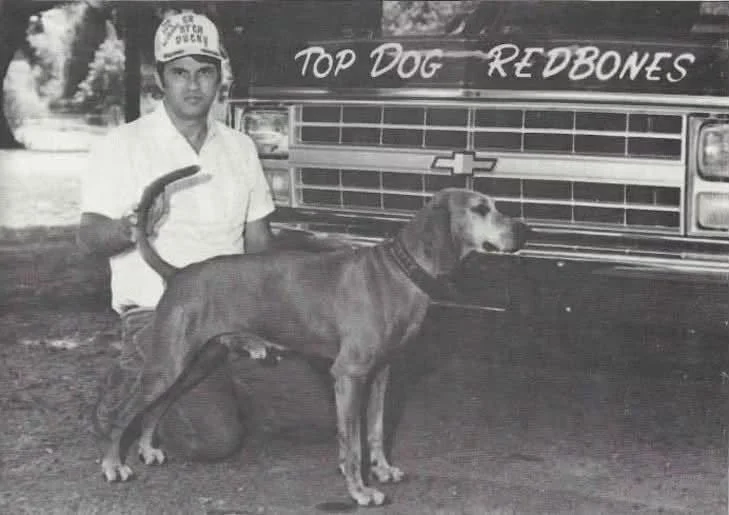A man kneels next to a large champion redbone coonhound in front of a vehicle with a sign that says "Top Dog Redbones."