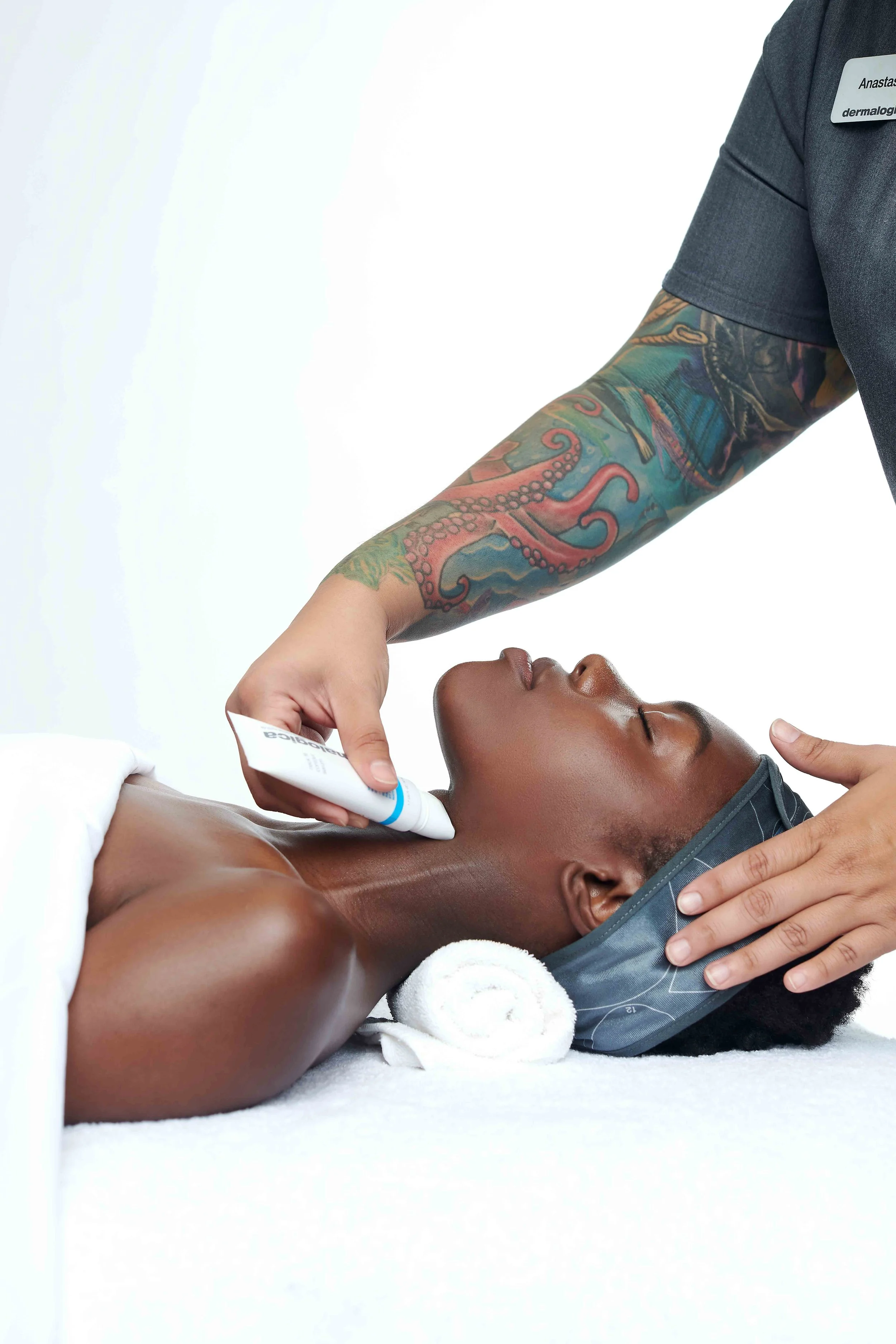 A person with a tattooed arm lying down and receiving a skincare or cosmetic treatment while a professional applies cream or lotion to their face.