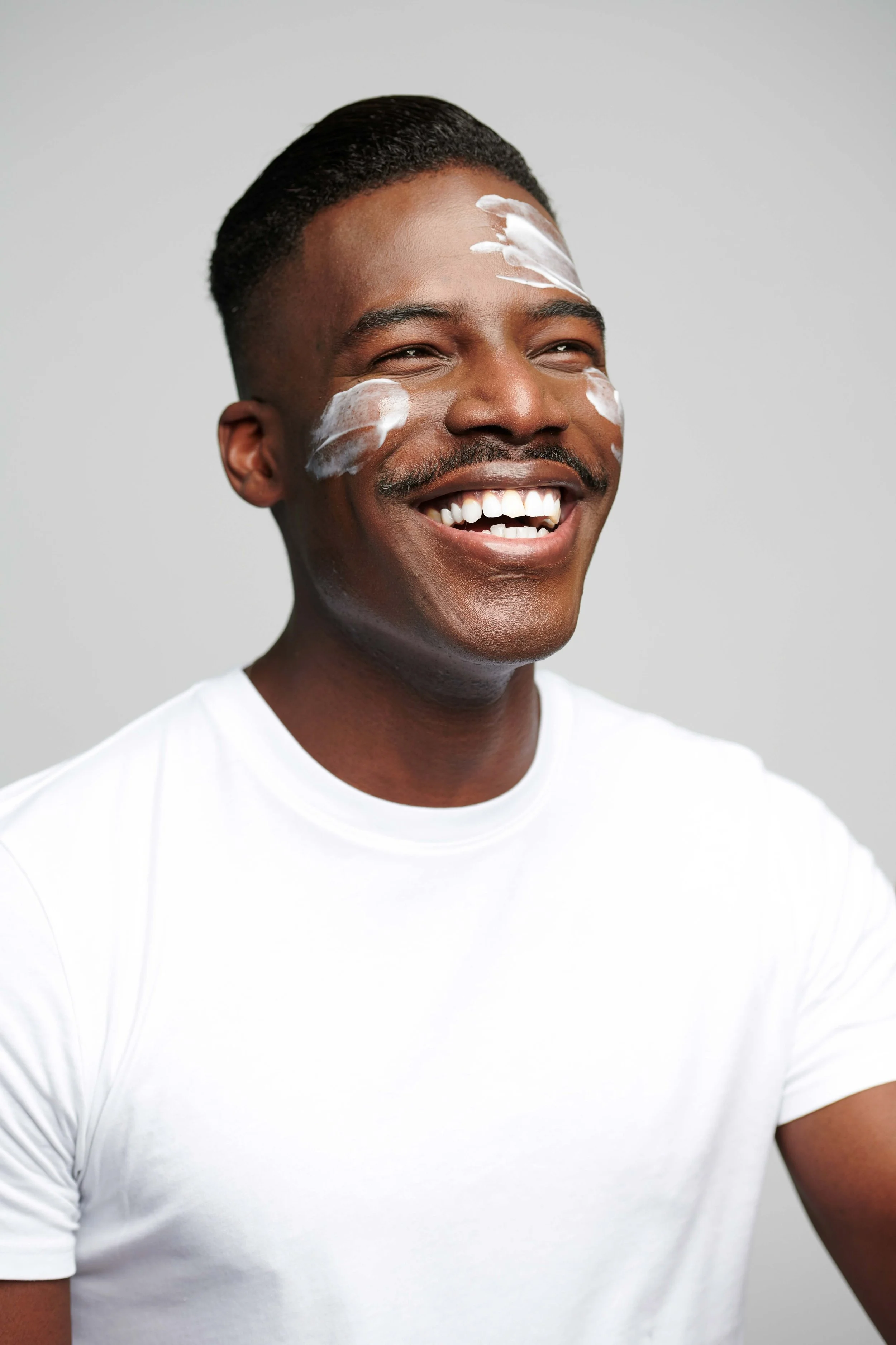 A happy Black man smiling with white sunscreen or moisturizer on his forehead and cheeks, wearing a plain white T-shirt, against a plain light gray background.