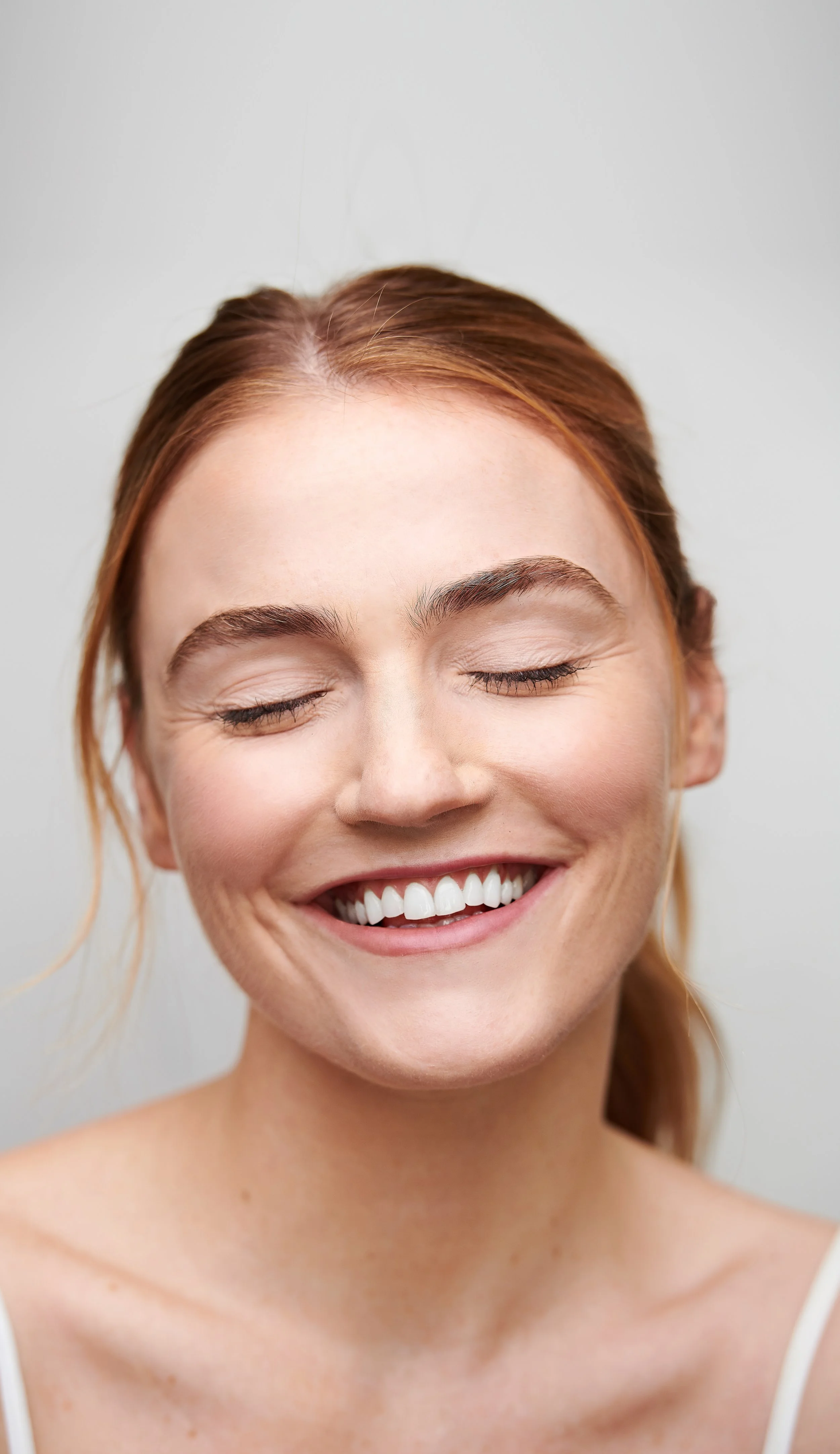 Close-up of a woman with red hair, smiling with eyes closed and showing white teeth, against a plain light background.