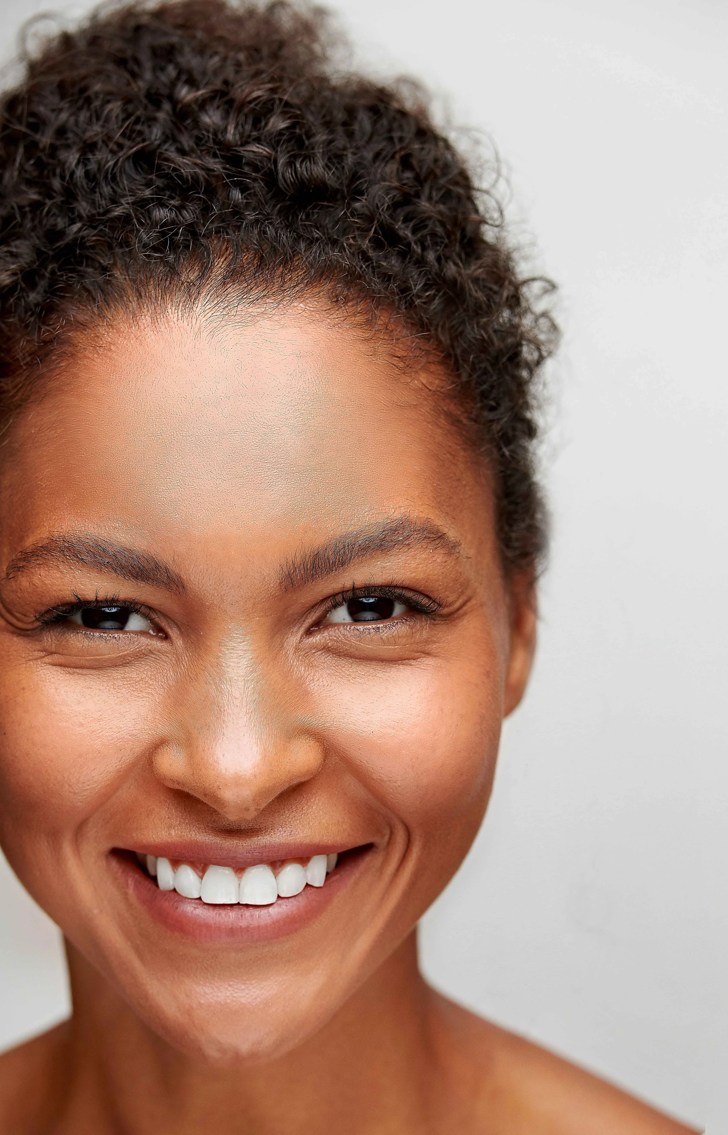 Close-up of a smiling woman with dark curly hair and clear skin, captured in a well-lit setting against a plain background.