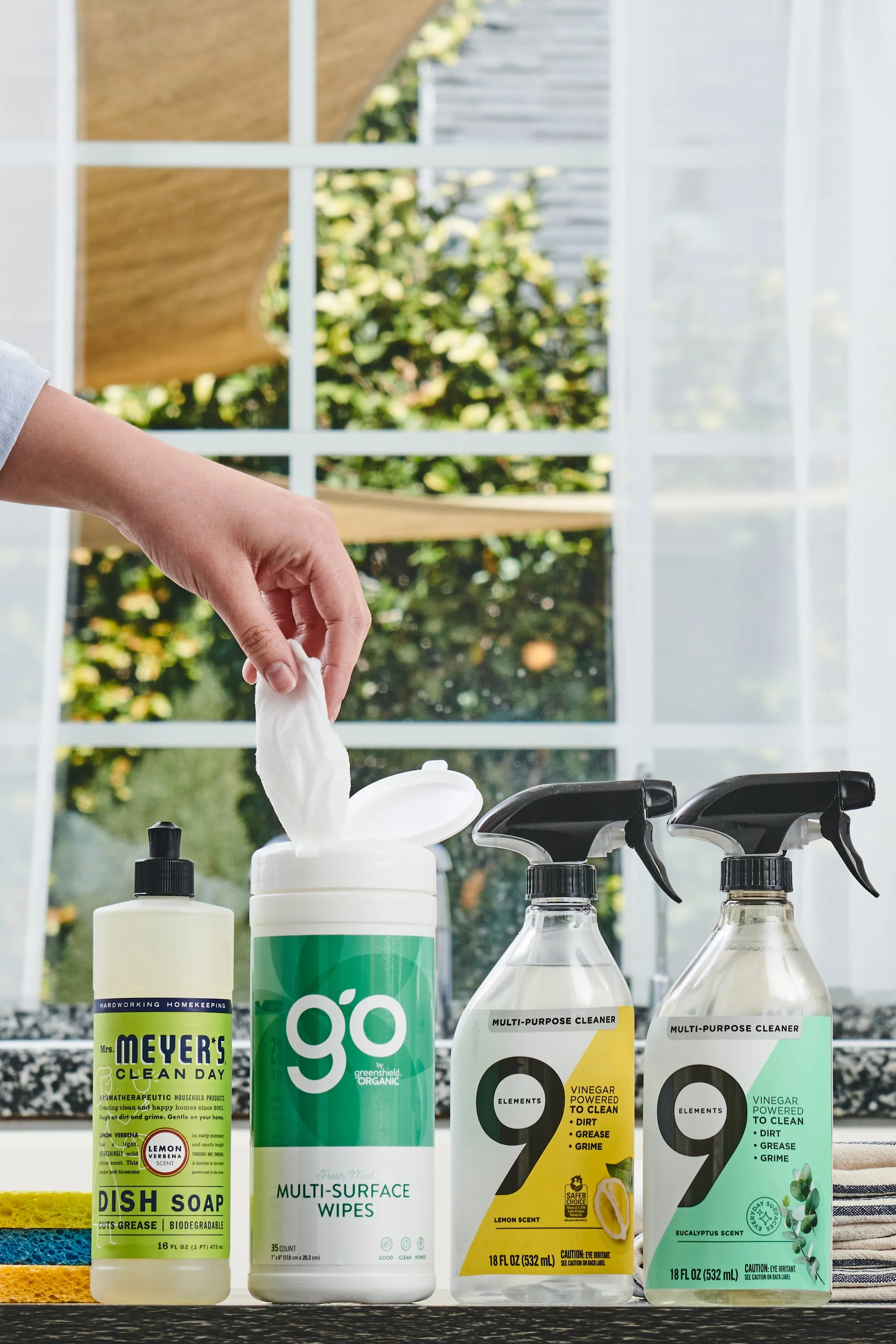 A collection of cleaning supplies on a windowsill, including dish soap, multi-surface wipes, and multi-purpose cleaners with spray nozzles.