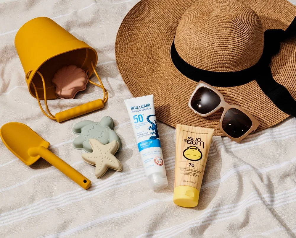 Beach items including a large sun hat, sunglasses, a yellow watering can, a small shovel, a toy turtle, a toy starfish, and sunscreen bottles on a striped fabric surface.