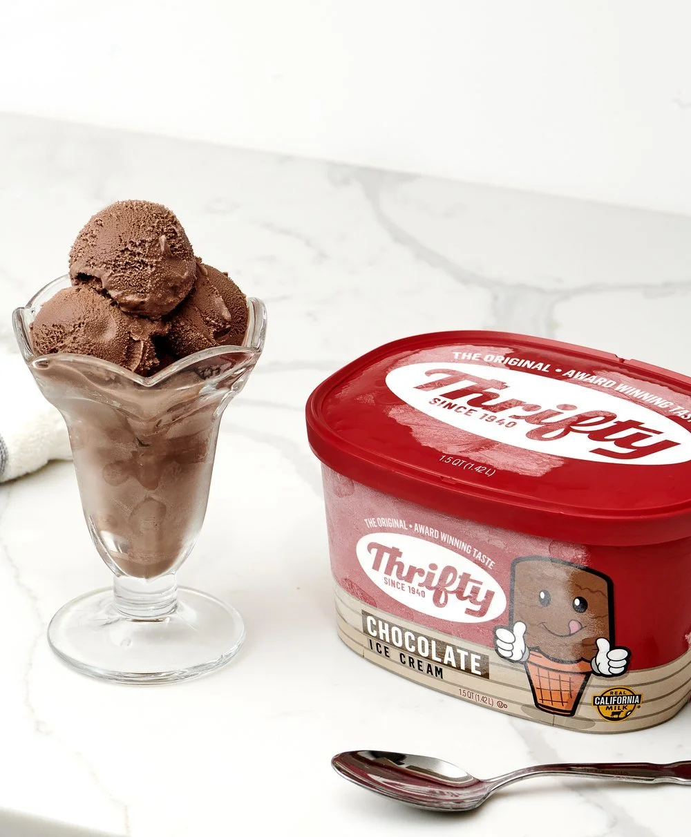 Chocolate ice cream scoops in a glass dish next to a tub of Thrify chocolate ice cream with a spoon on a white marble surface.