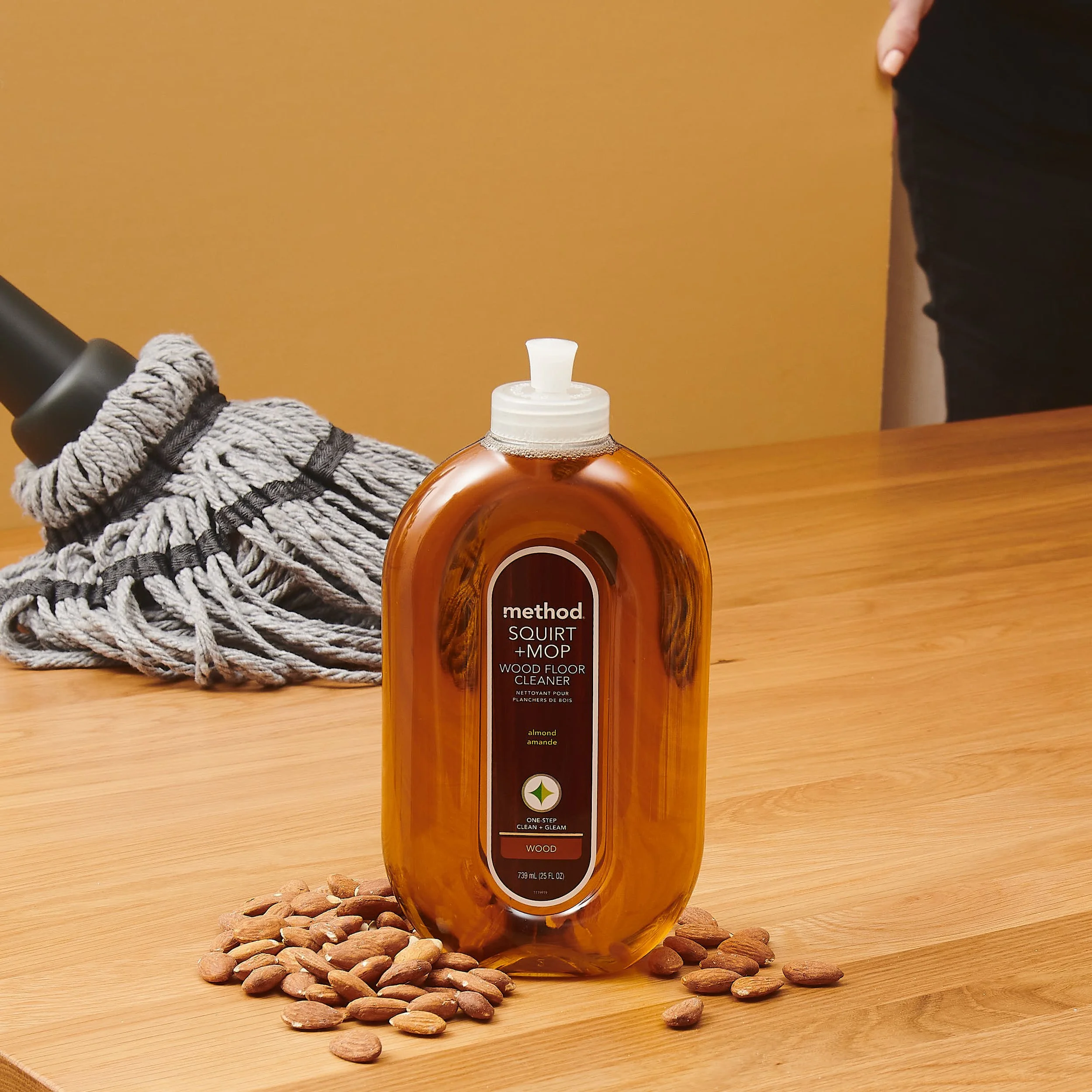 A clear orange bottle of Method Squirt + Mop Wood Floor Cleaner on a wooden surface, with a pile of almonds and a rope in the background, and a person in black shorts to the right.