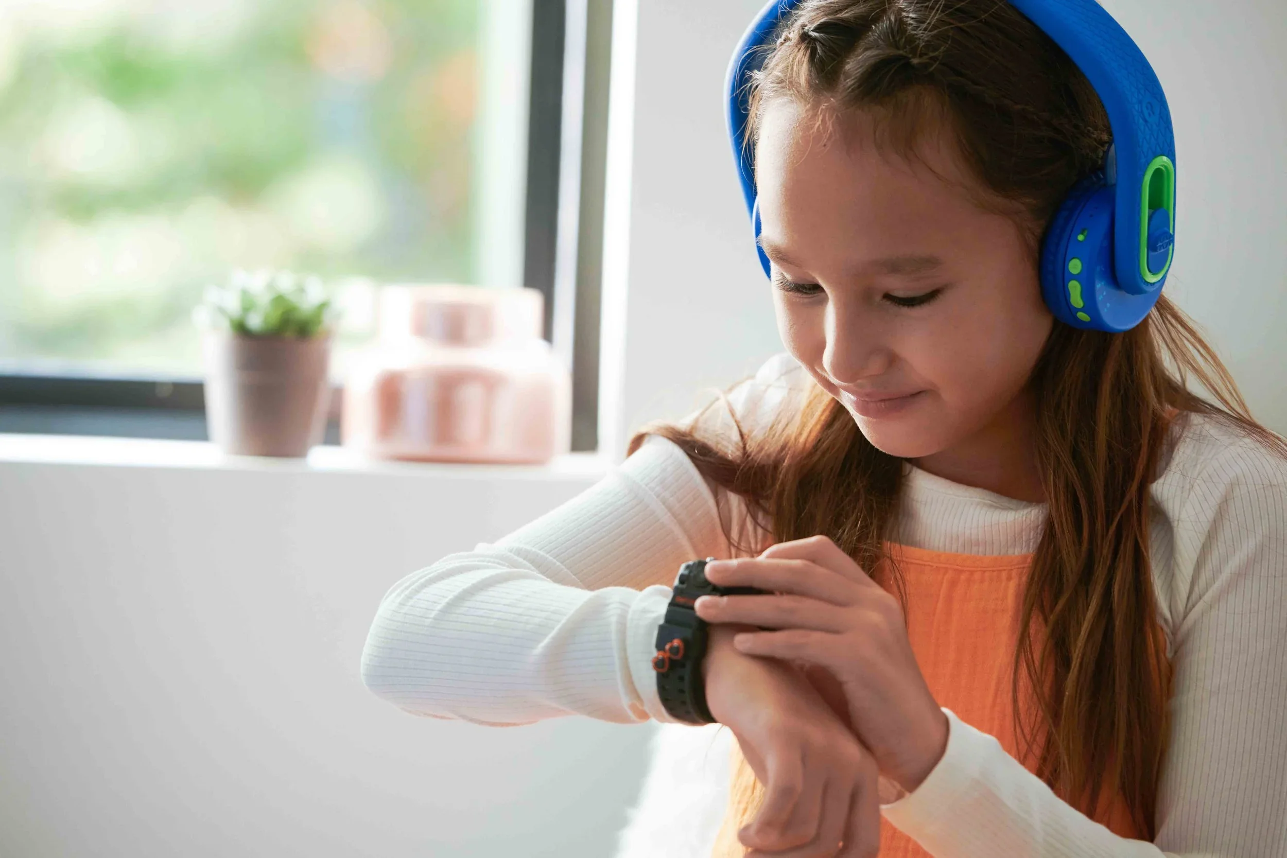 Young girl with long red hair wearing a blue and green headset, looking at her smartwatch, in a brightly lit room with a window and potted plant in the background.