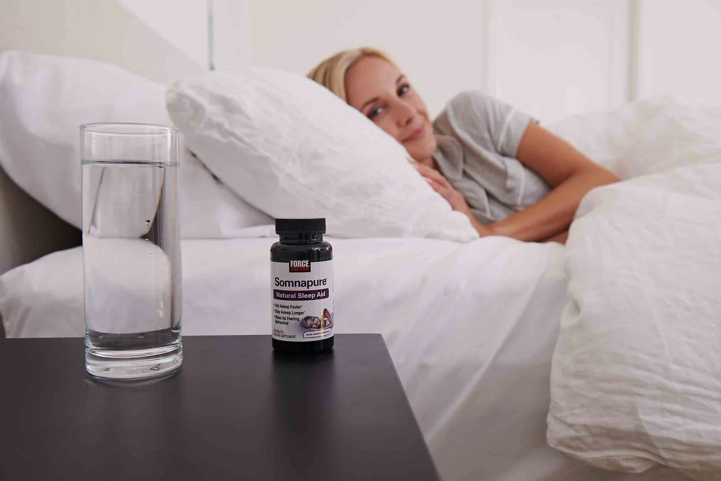 A woman lying in bed with a glass of water on a nightstand and a bottle of herbal sleep aid supplement labeled 'Sommature' in the foreground.