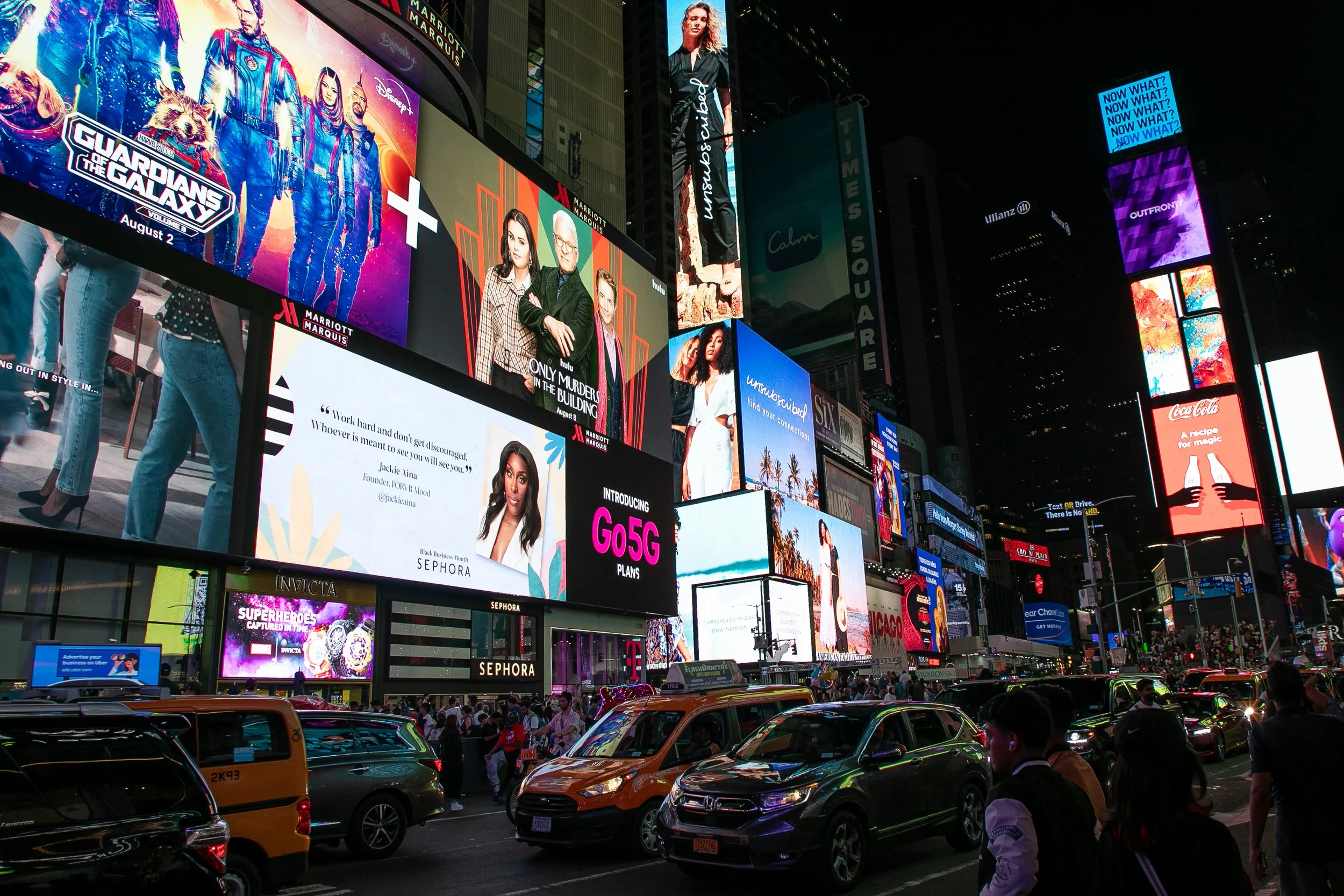 Nighttime view of Times Square in New York City with illuminated digital billboards and posters, and a busy street filled with cars and pedestrians.