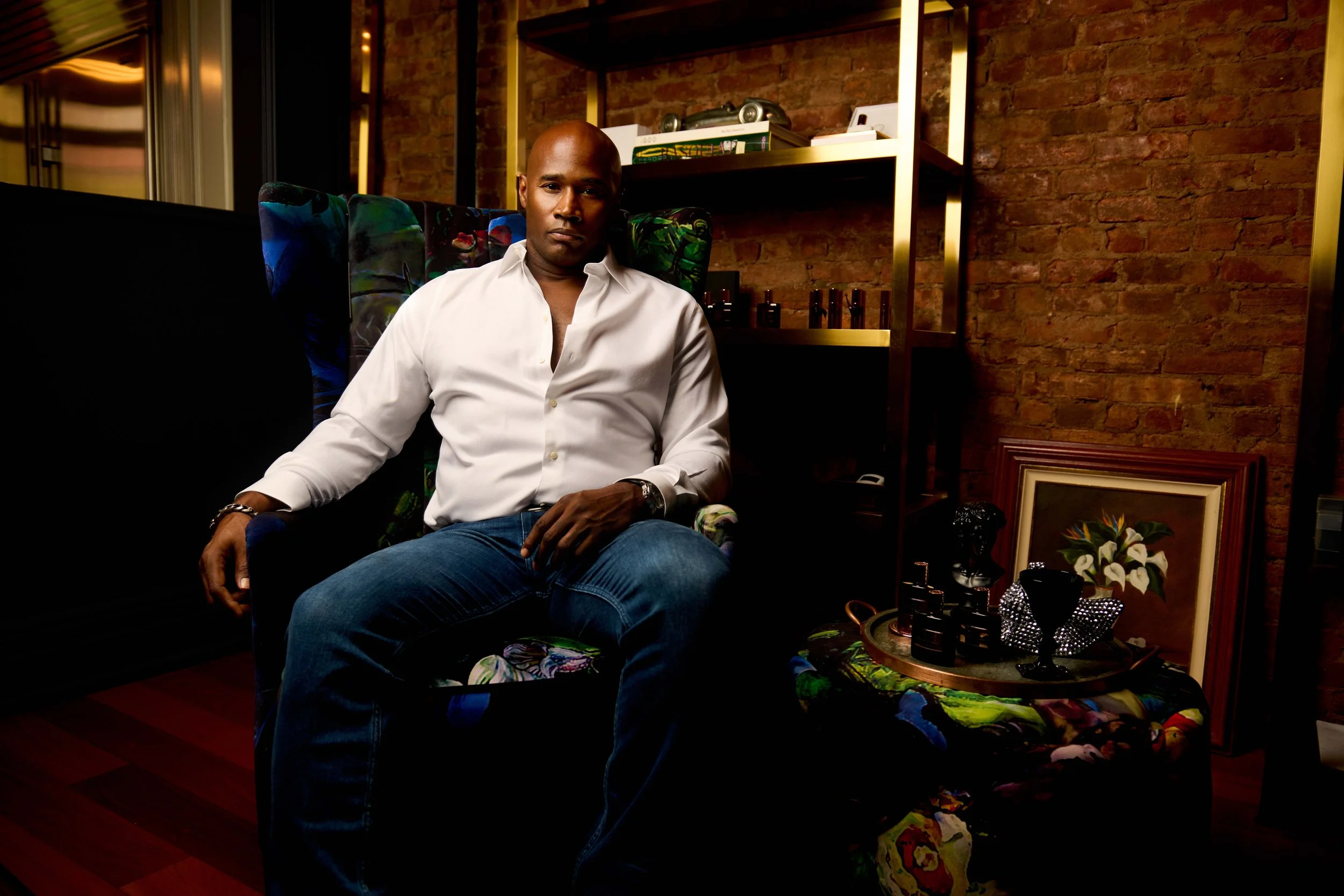 A man with dark skin and a shaved head sitting on a colorful floral armchair in a room with a brick wall background, next to a small round table with decorative objects and a framed picture leaning against the wall.