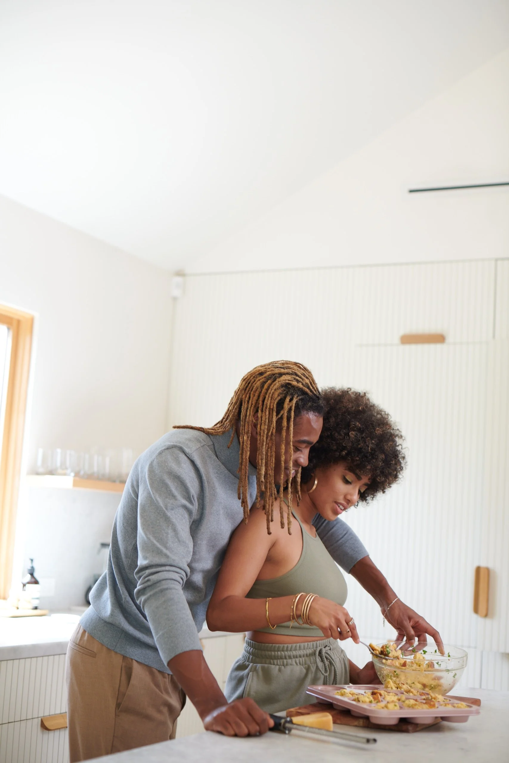 A man with dreadlocks and a woman with curly hair preparing food together in a bright kitchen.