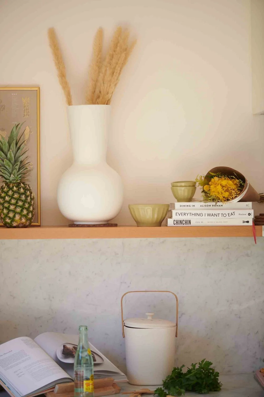 Decorative shelf with a white vase holding dried pampas grass, a framed pineapple print, stacked books, green bowls, a flower bouquet wrapped in black paper, a glass bottle, and a white container with a handle, with an open magazine and green foliage