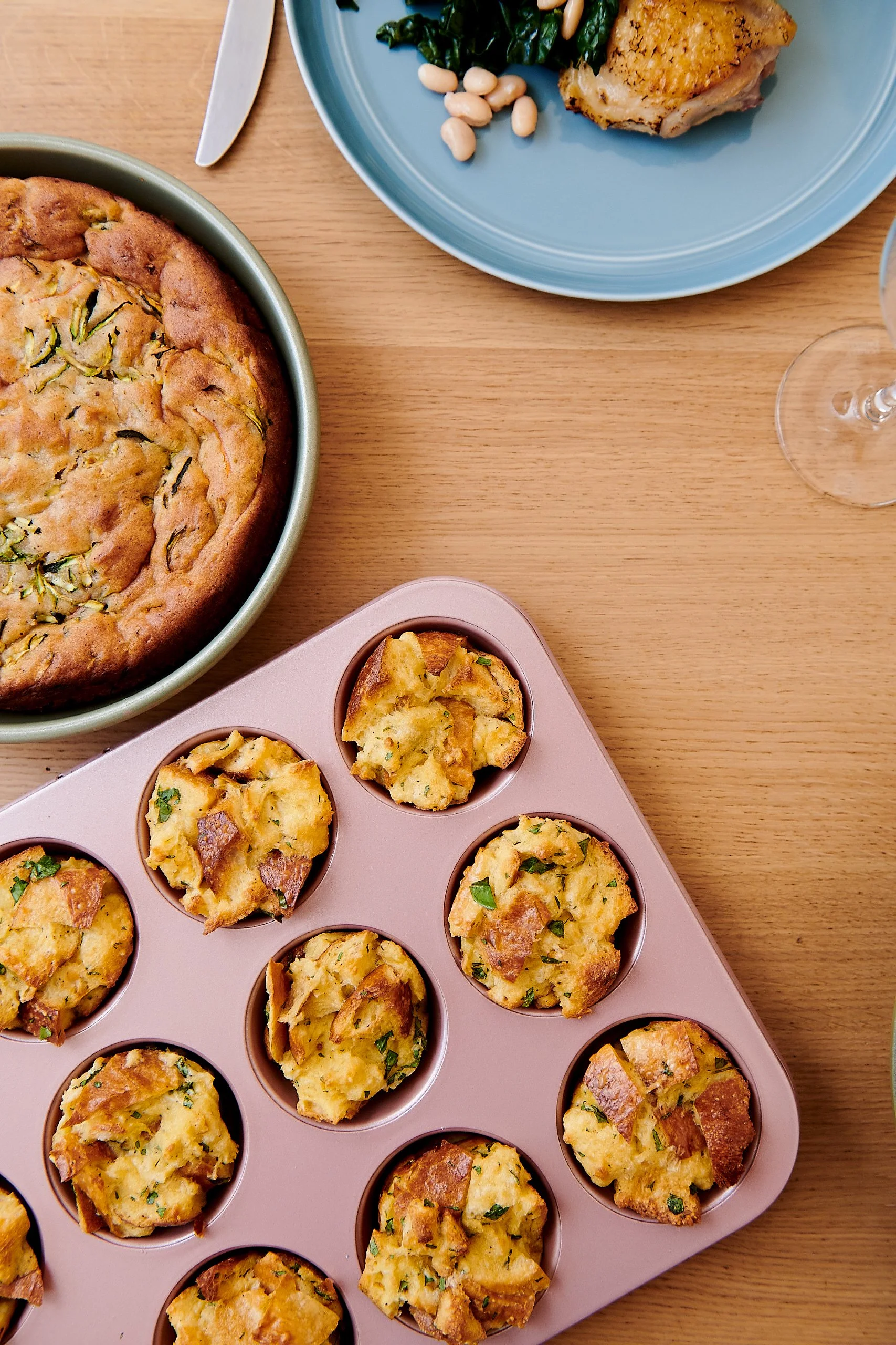 A table with a large round zucchini frittata in a green dish, a blue plate with a piece of breaded chicken, cooked spinach, white beans, and a glass of wine. A pink muffin tray has baked egg muffins with herbs and pieces of meat.