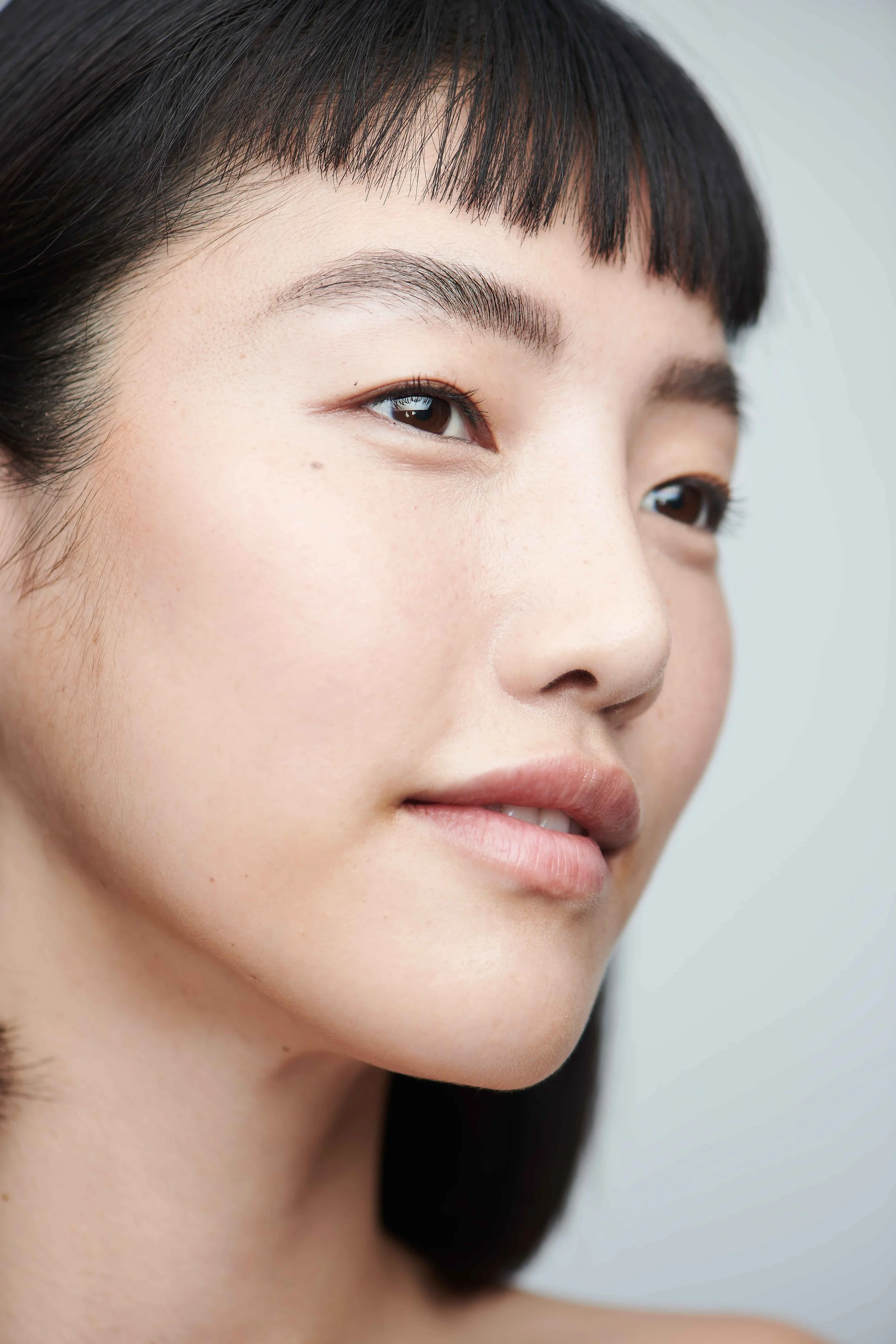 Clean skincare beauty portrait of a woman with a blunt fringe and bare makeup on a pale grey backdrop.