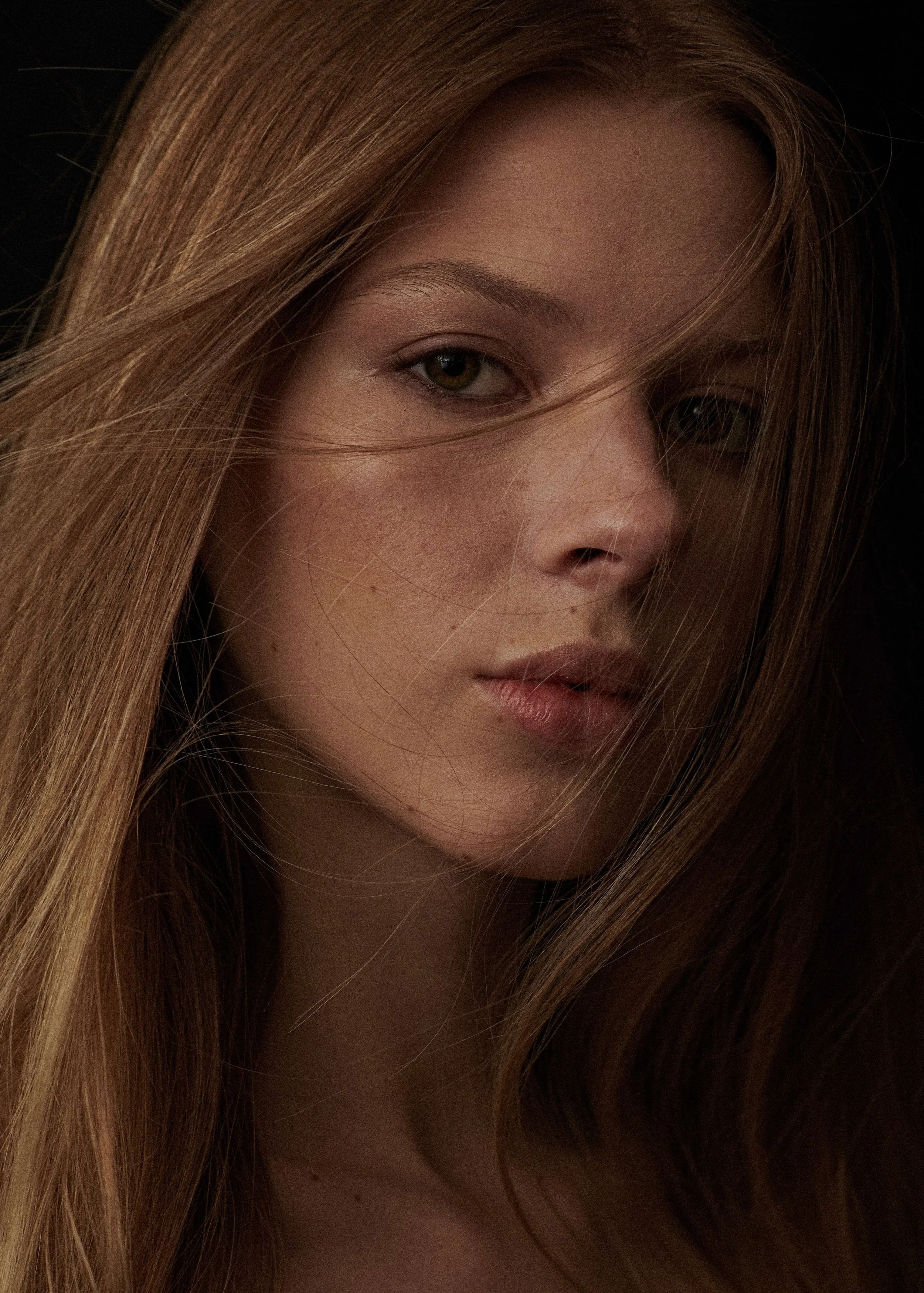 Low-key beauty portrait of a woman with long auburn hair and freckles, hair falling across her face.