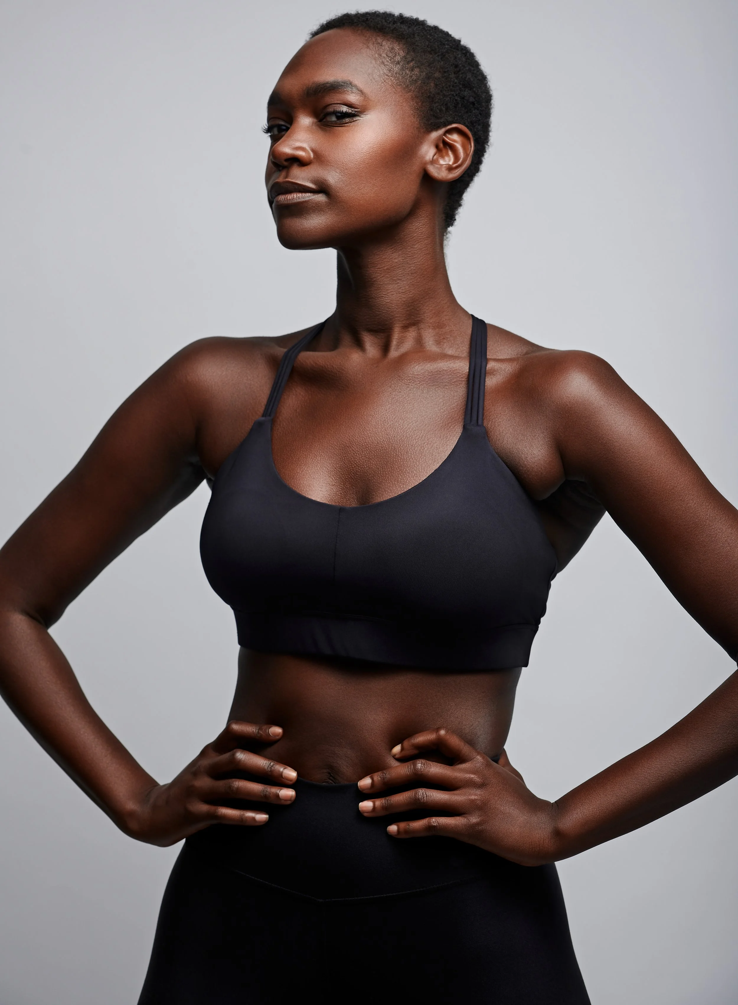 Studio beauty and body shot of a woman in black activewear, hands on hips, on a grey backdrop.