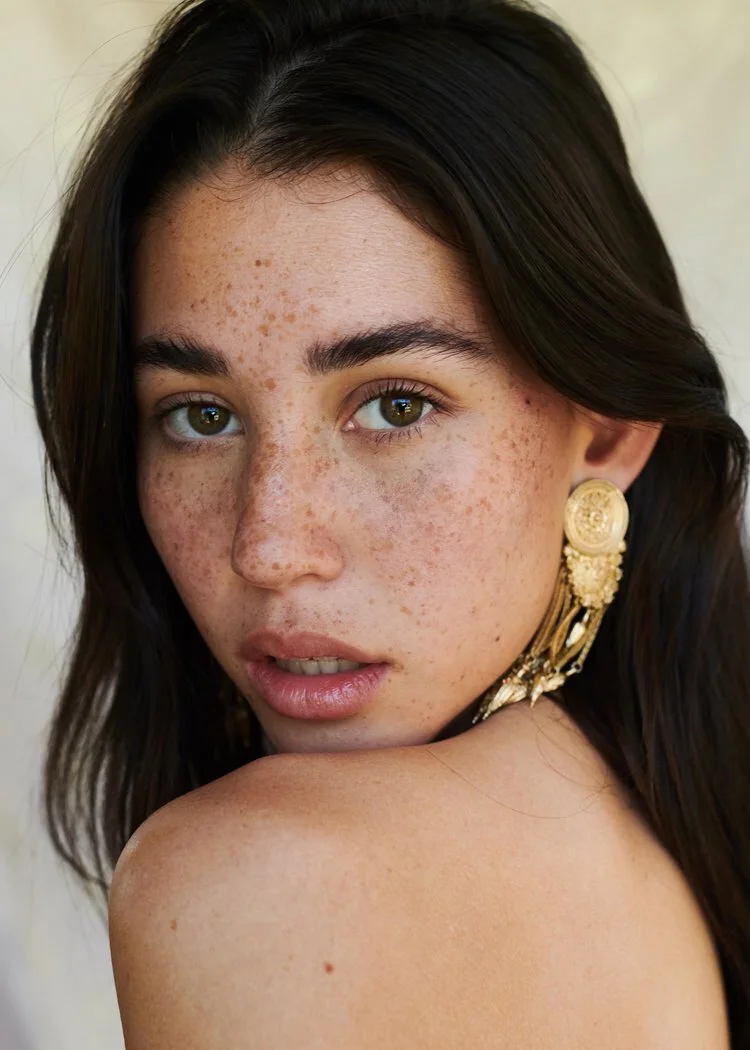 Beauty portrait of a woman with freckles and a gold statement earring looking over her bare shoulder.