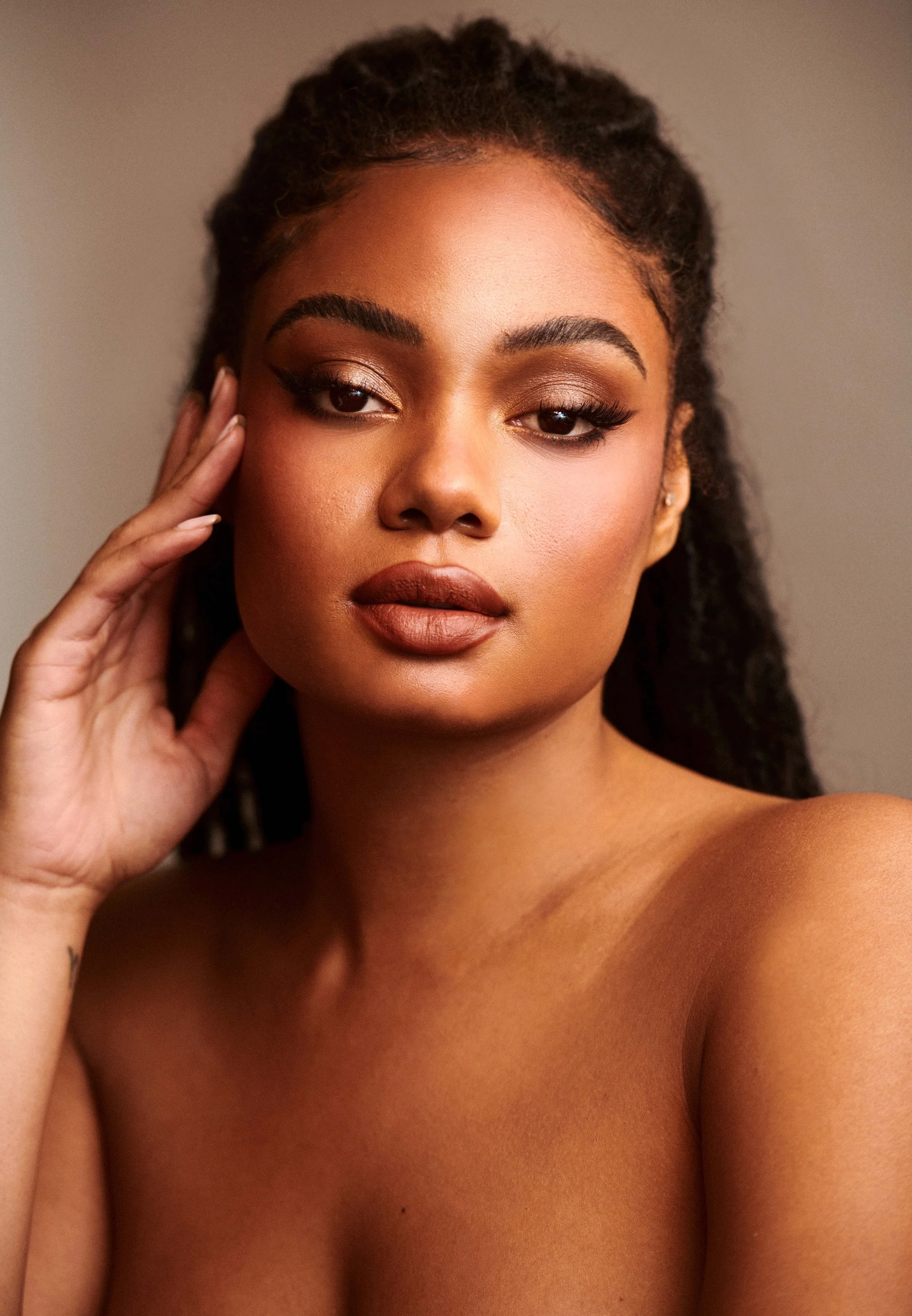 Glowing beauty portrait of a woman with a hand to her temple, warm neutral makeup on a tan backdrop.