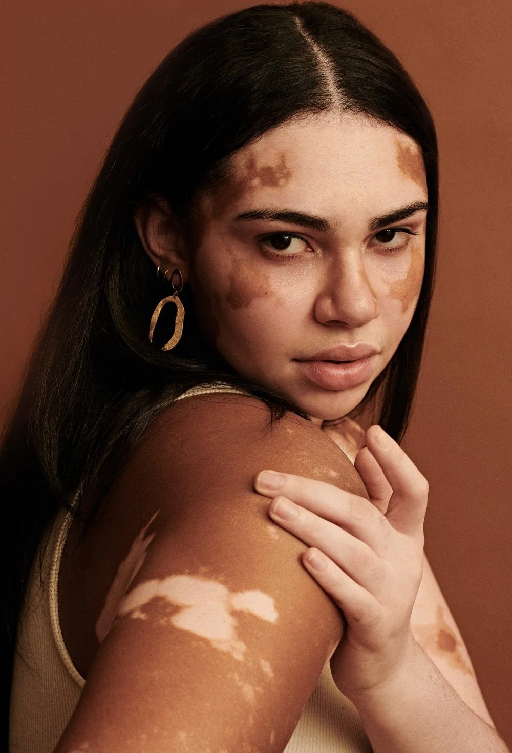 Beauty portrait of a woman looking over her shoulder with a gold hoop earring, shot against a warm terracotta background.