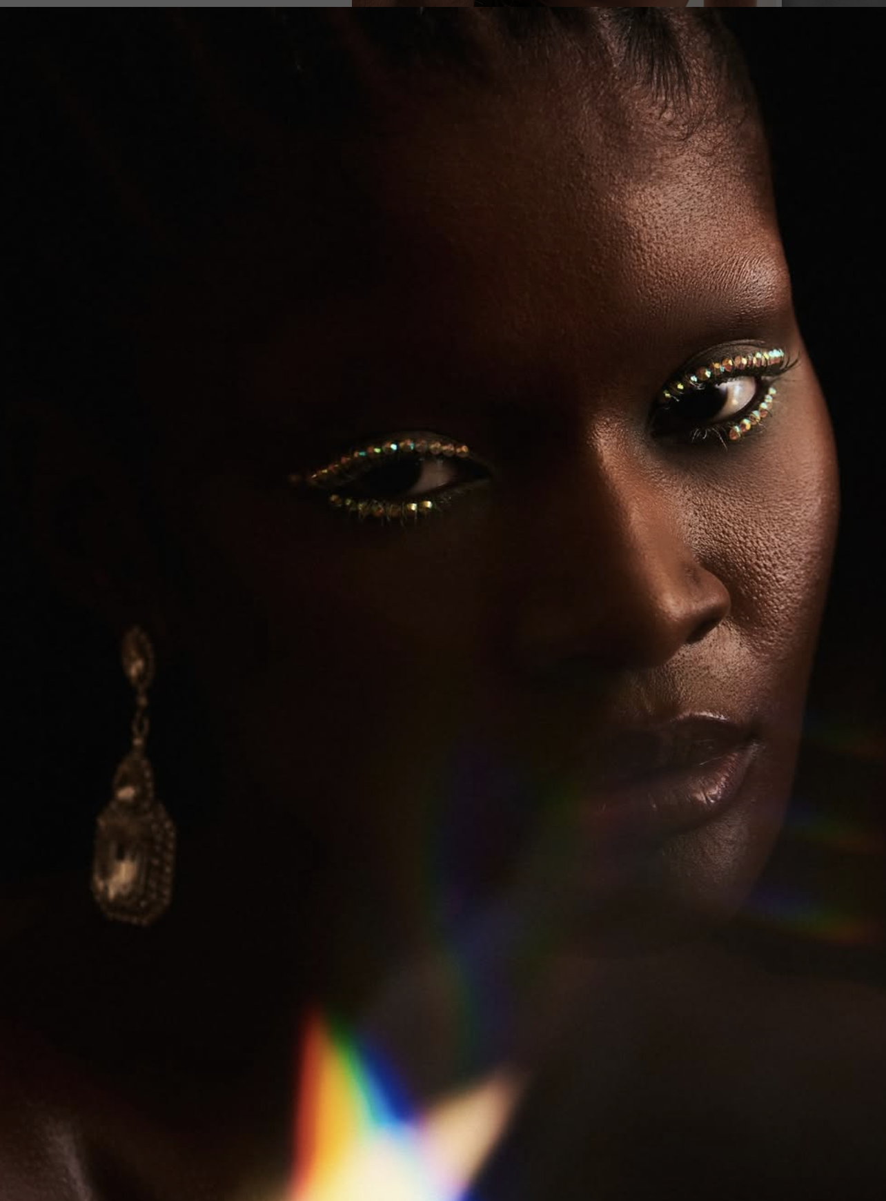 Low-key beauty portrait of a woman in gold glitter eye makeup and a drop earring, prism flare across the frame.