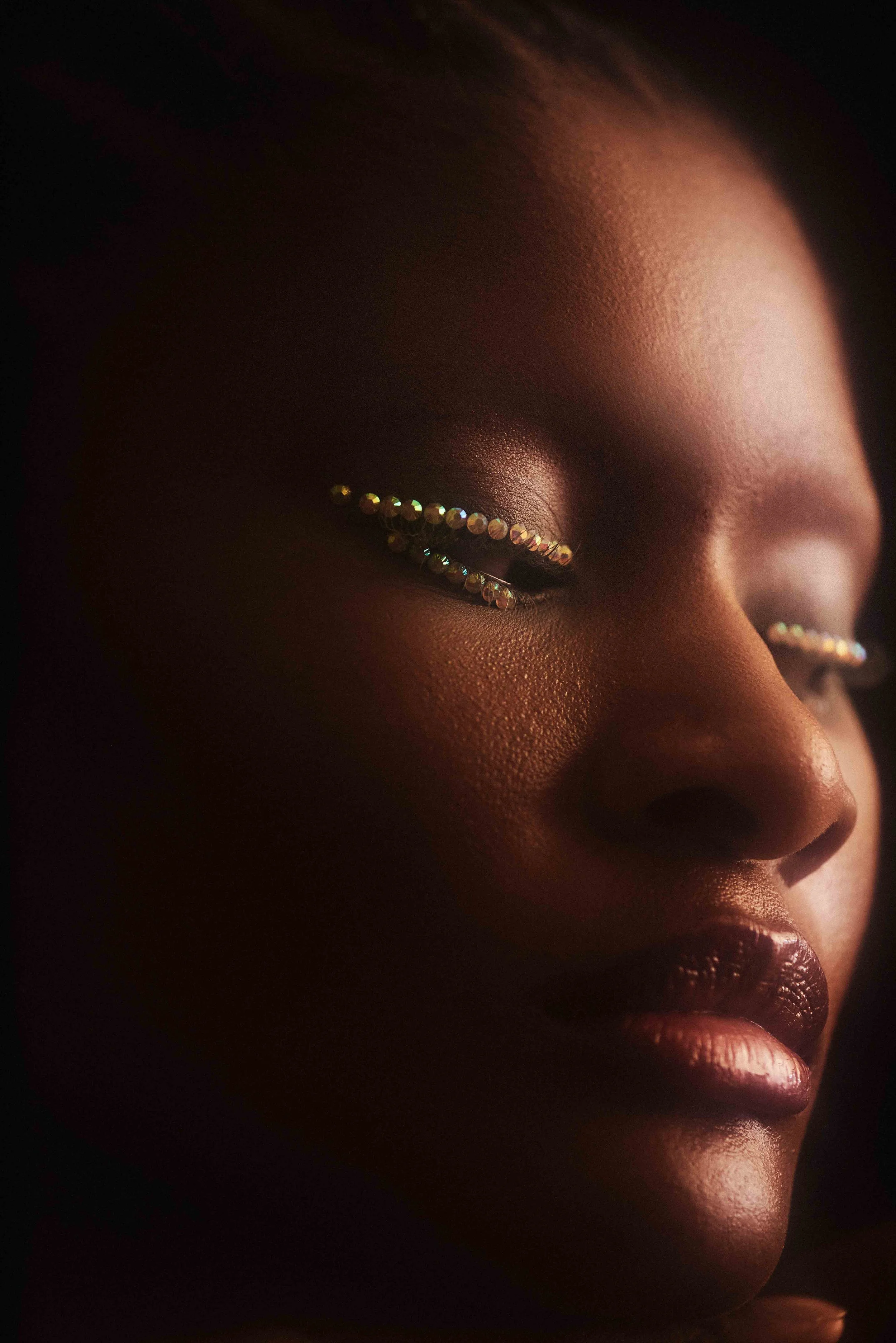 Macro beauty frame of gold beaded lashes on a closed eye, warm directional light on deep skin.