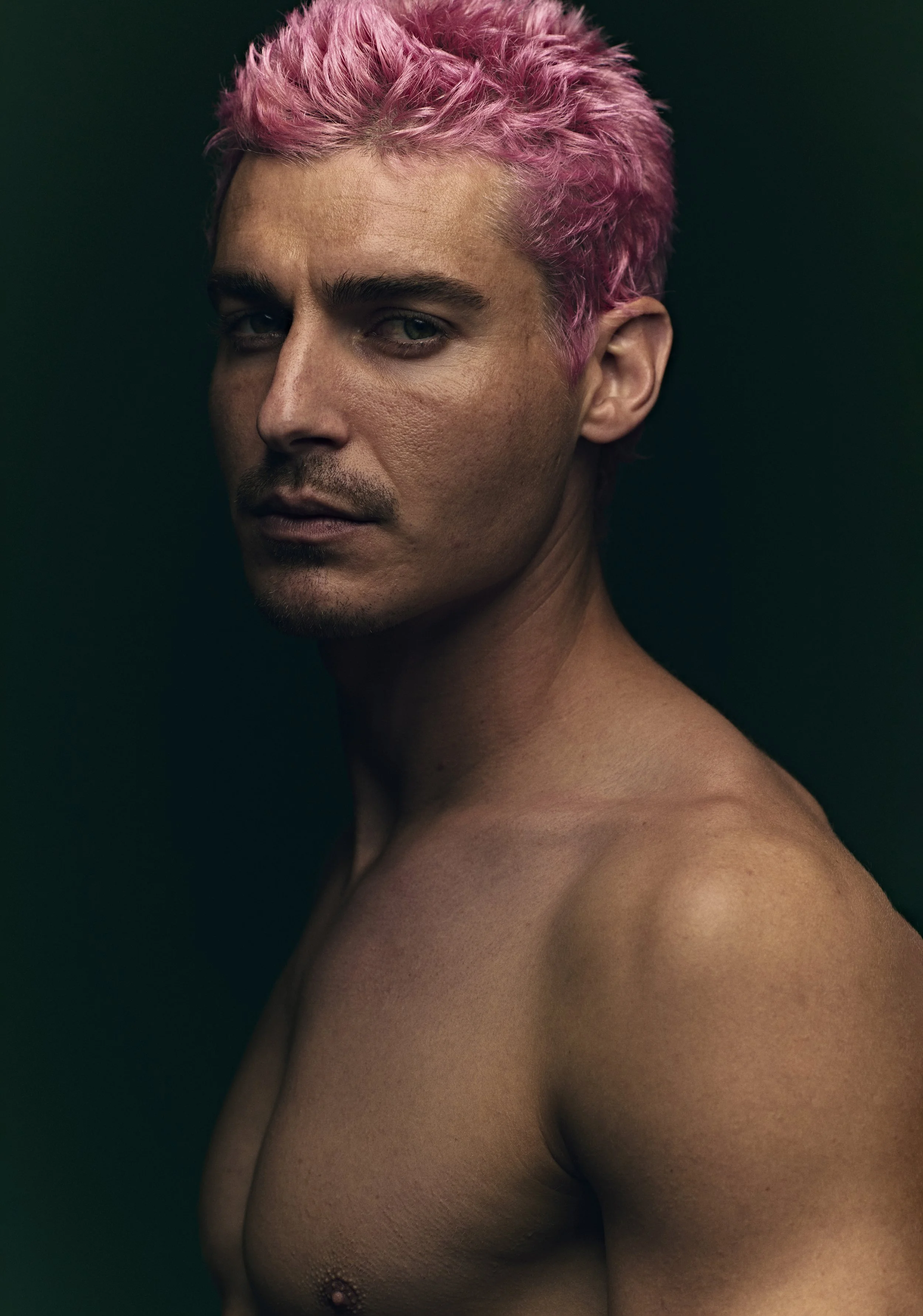 Beauty portrait of a man with bleached pink hair and a moustache, bare shoulders against a dark green backdrop.