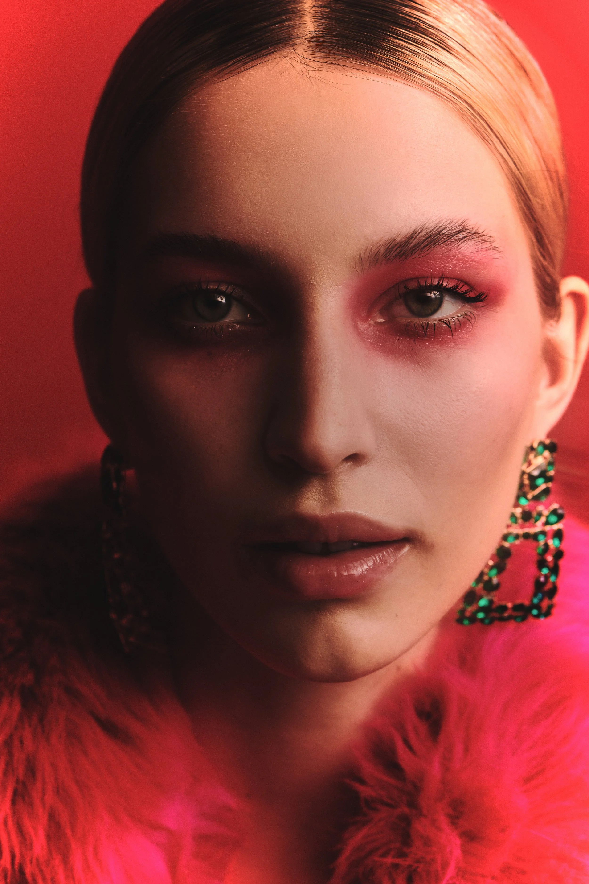 Colour beauty editorial: a woman in pink blush-toned eye makeup and jewelled earrings against a red gel background.