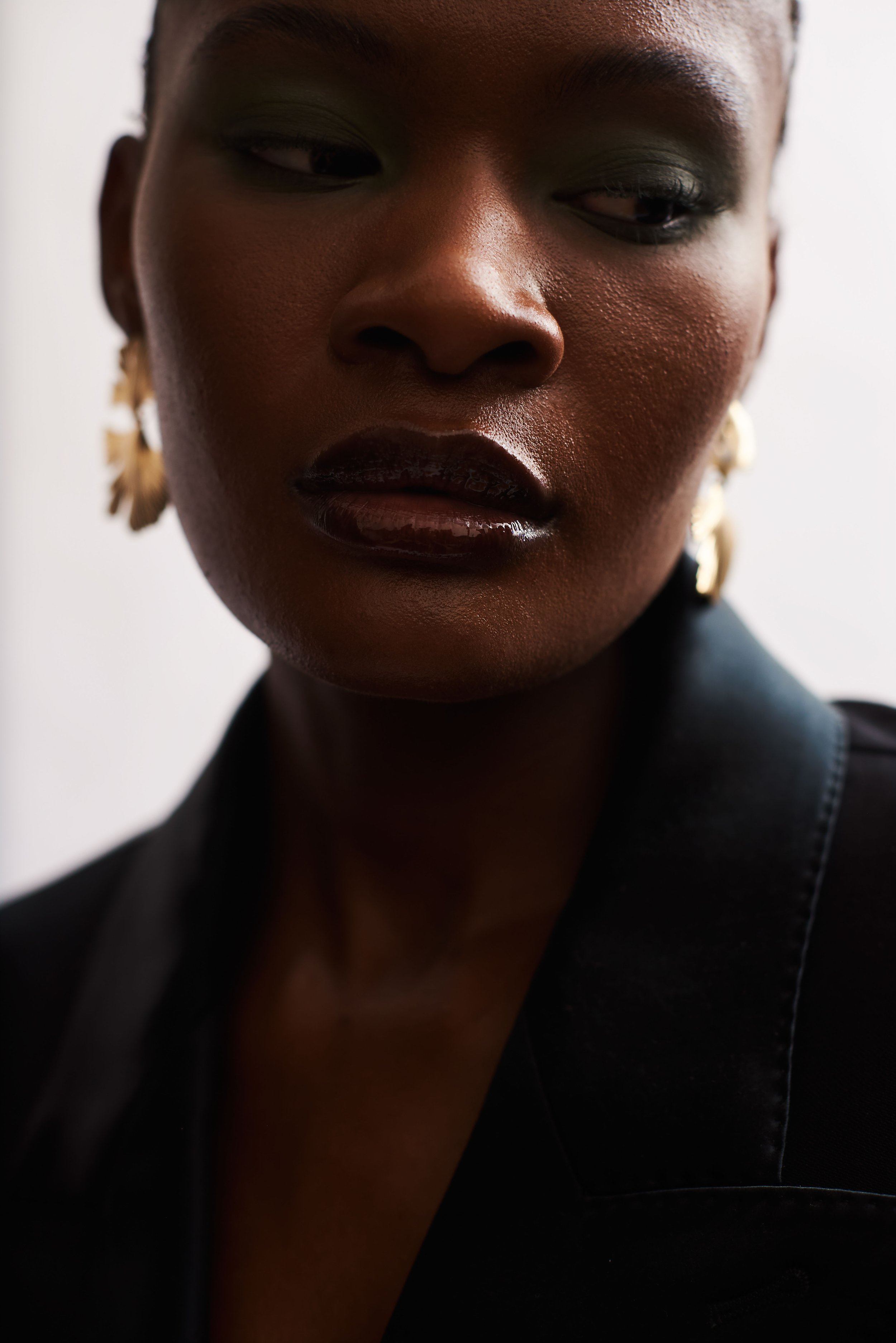Beauty portrait of a woman in green eyeshadow, glossed lips and gold hoop earrings, wearing a black blazer.