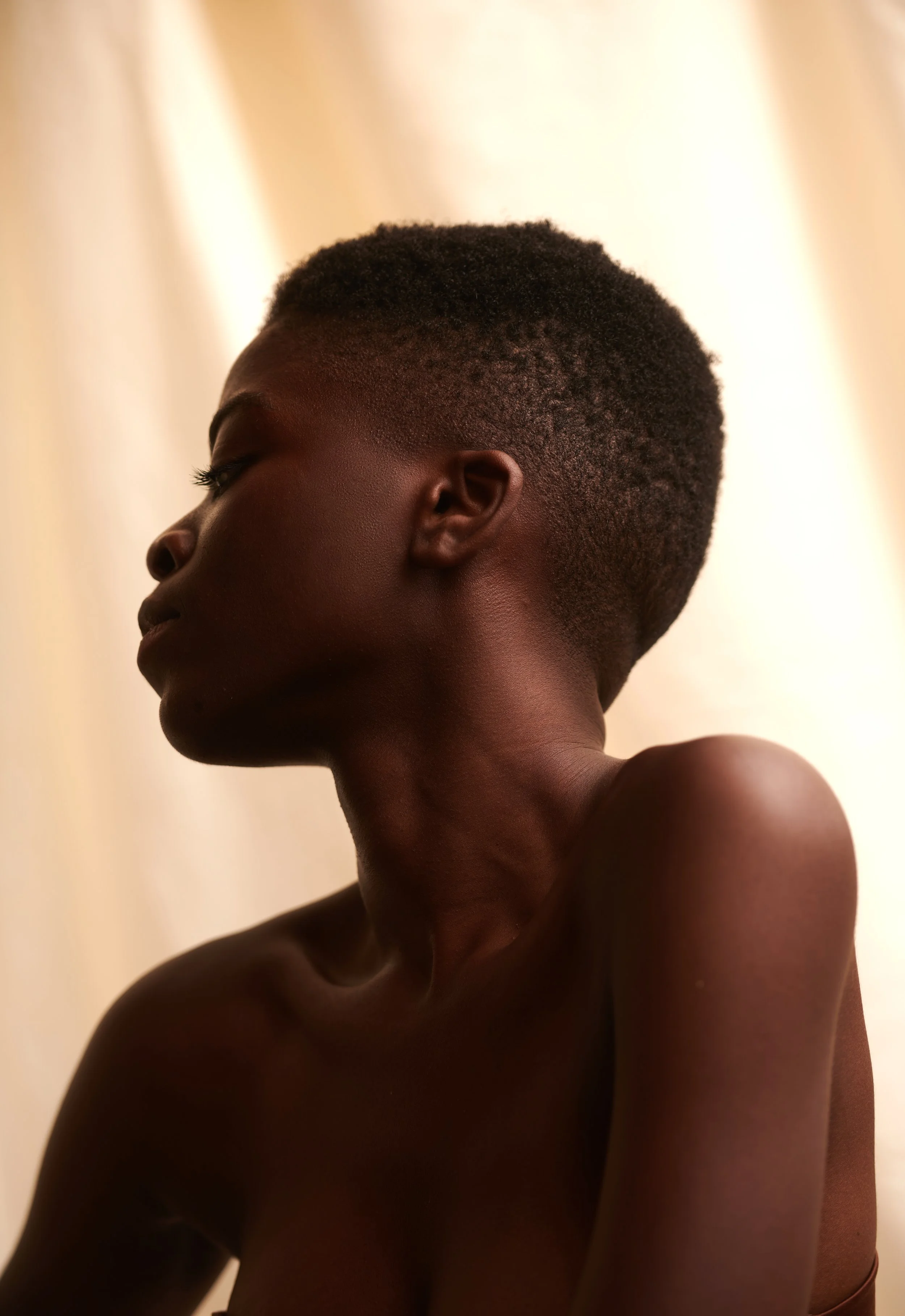 Beauty profile portrait of a woman with a close crop and bare shoulders, warm window light on draped fabric.