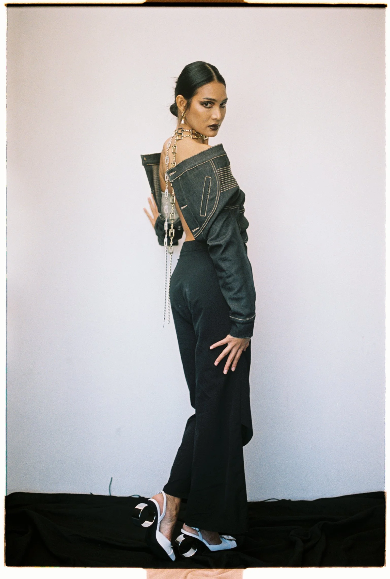 Full-length studio test shoot of a model in an off-shoulder corset top, black trousers and chain jewellery on a grey backdrop.