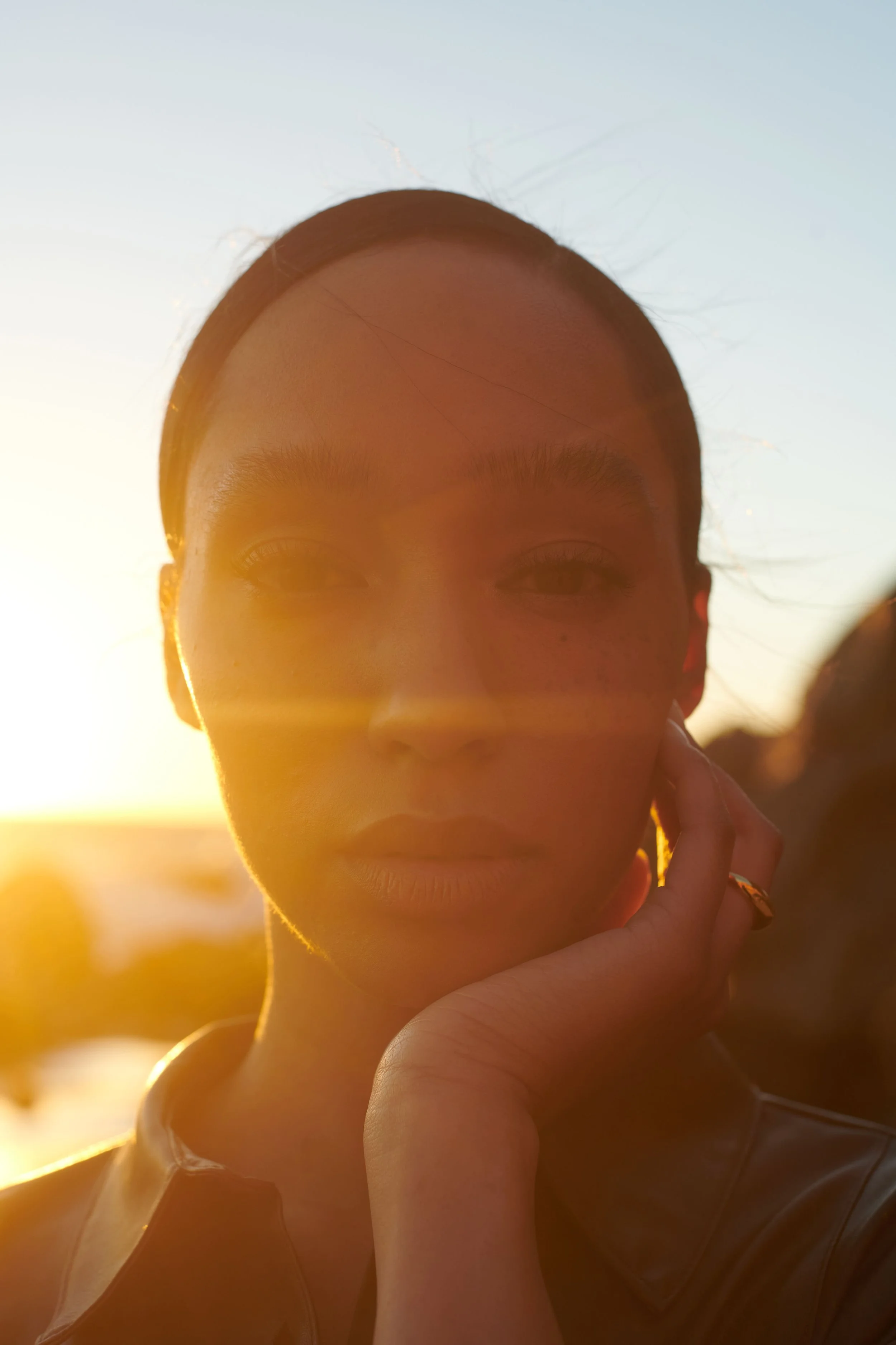Golden-hour beauty test portrait of a woman with her hand to her face, sun flare crossing the frame.