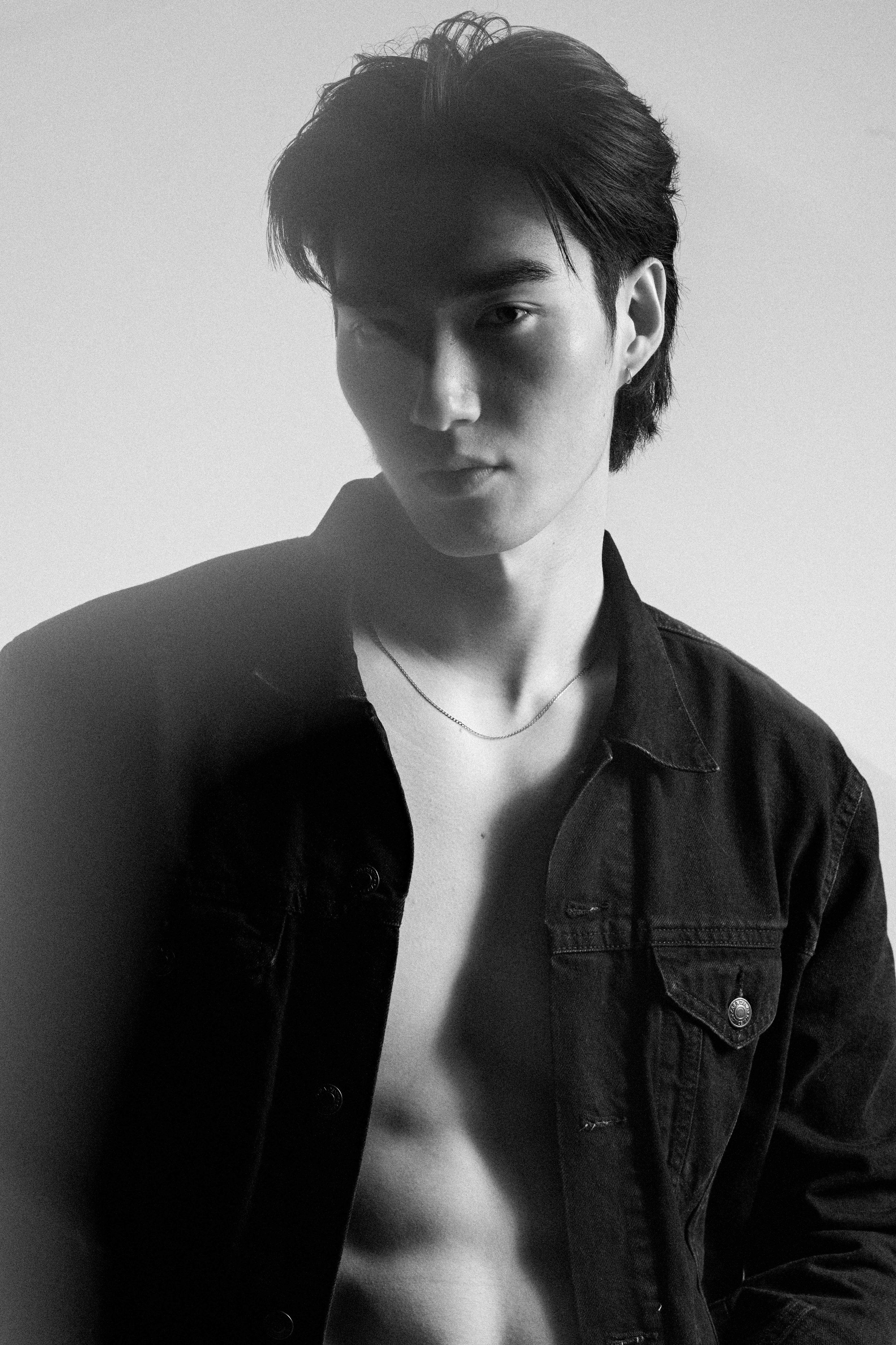 Black-and-white fashion test portrait of a man in an open dark denim shirt and fine chain, lit against a pale wall.