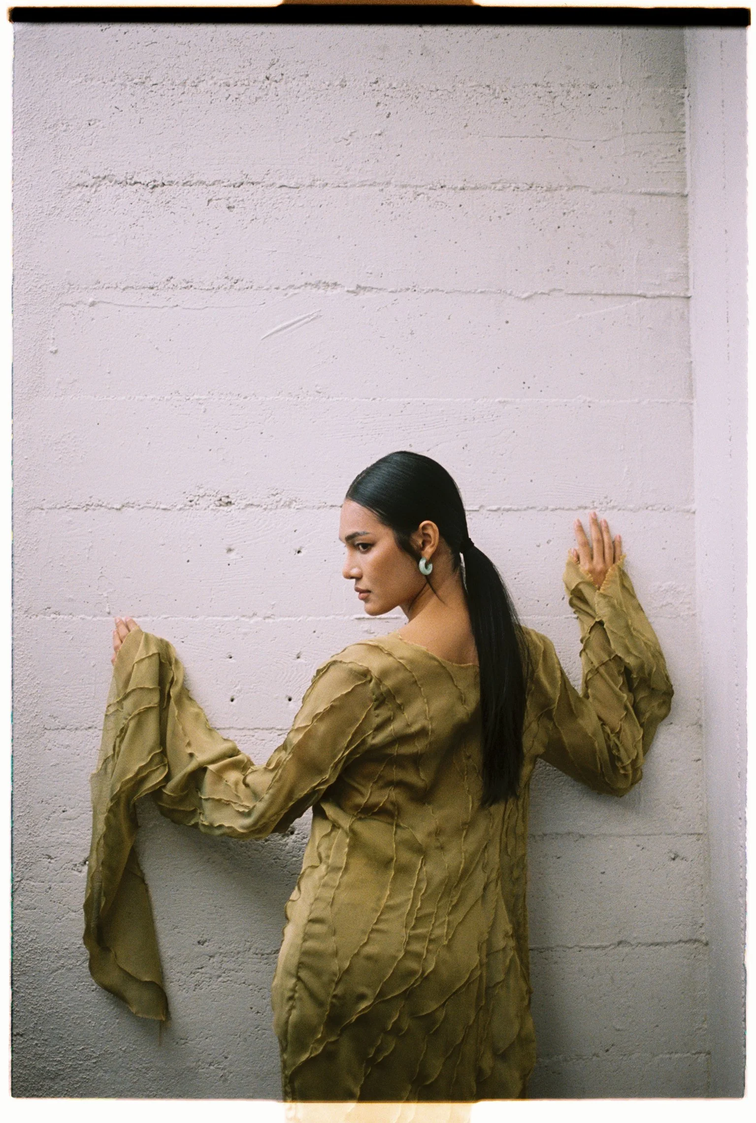 Location test shoot of a woman in an olive draped dress against a painted brick wall in daylight.