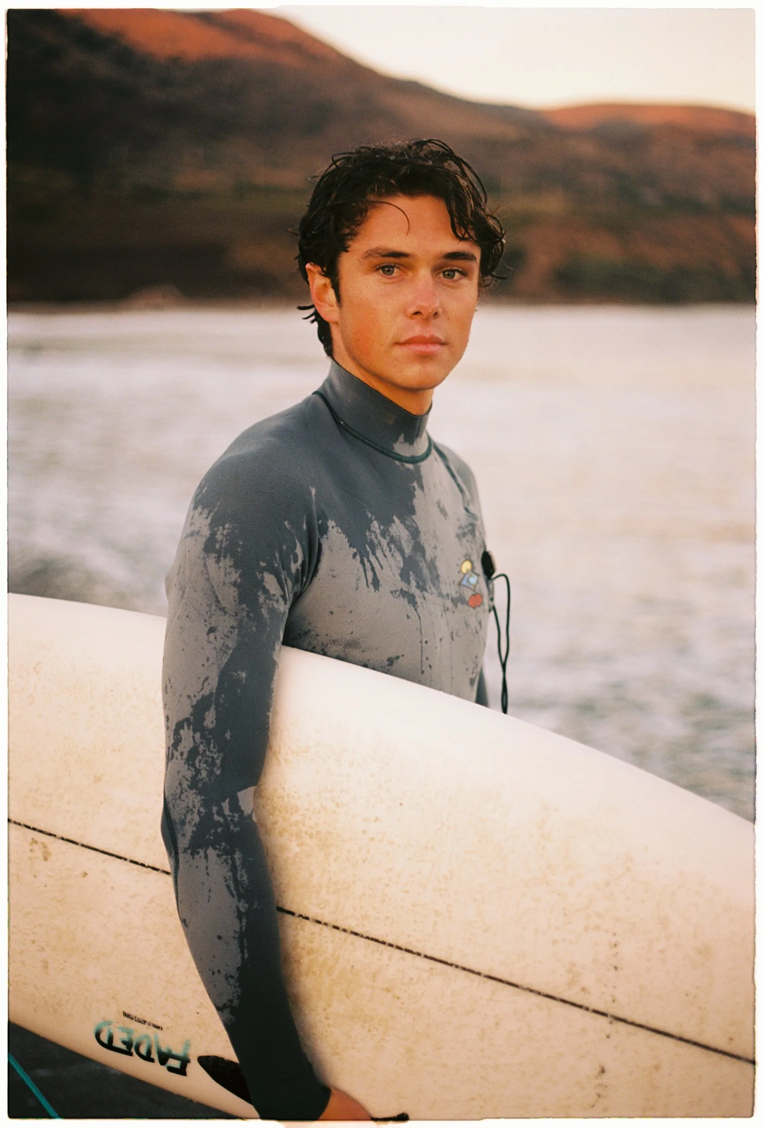 Coastal test shoot of a young man in a wetsuit carrying a surfboard, overcast light on the California shoreline.