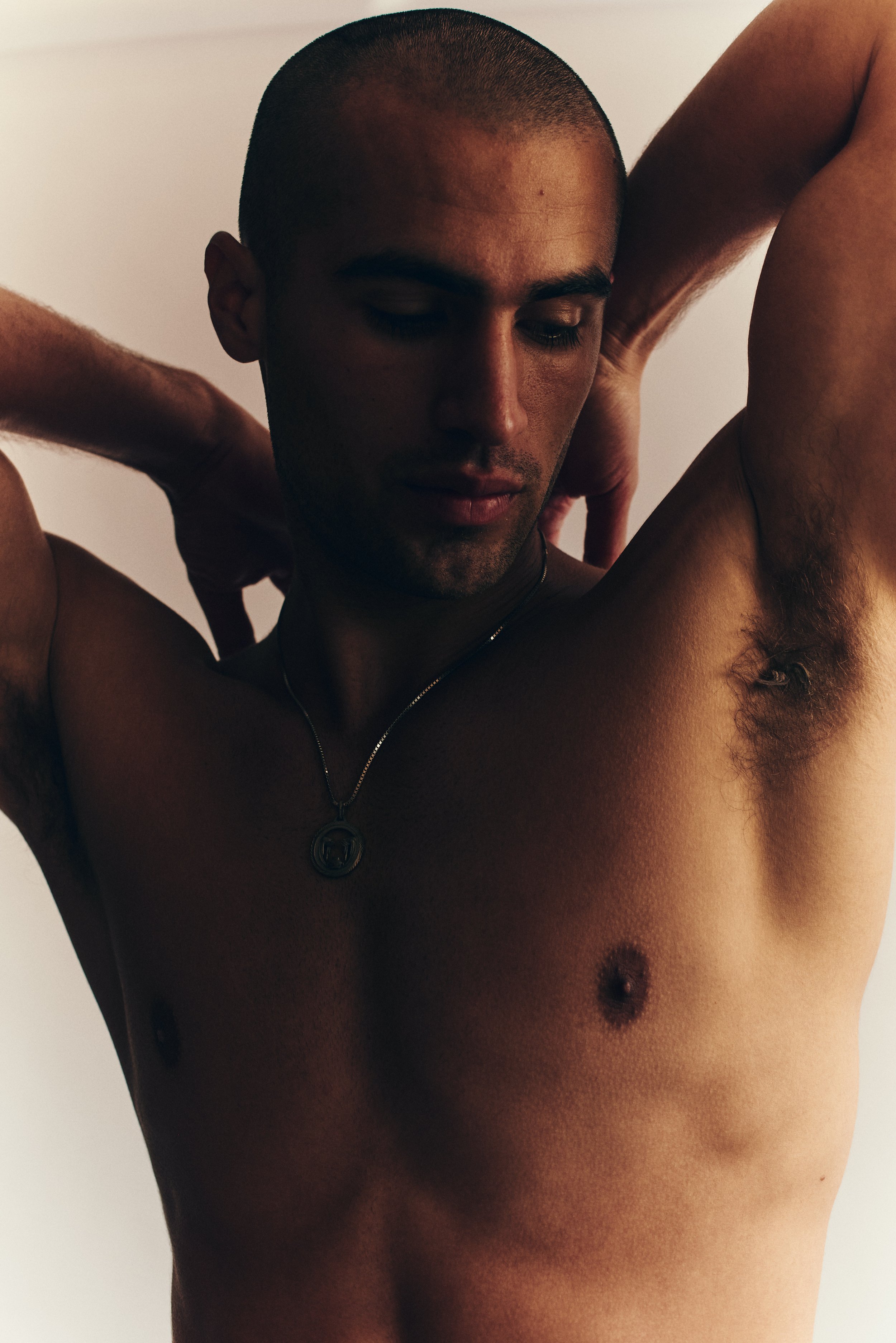 Warm-toned test portrait of a shirtless man with a shaved head, arms raised behind his head, soft directional light.