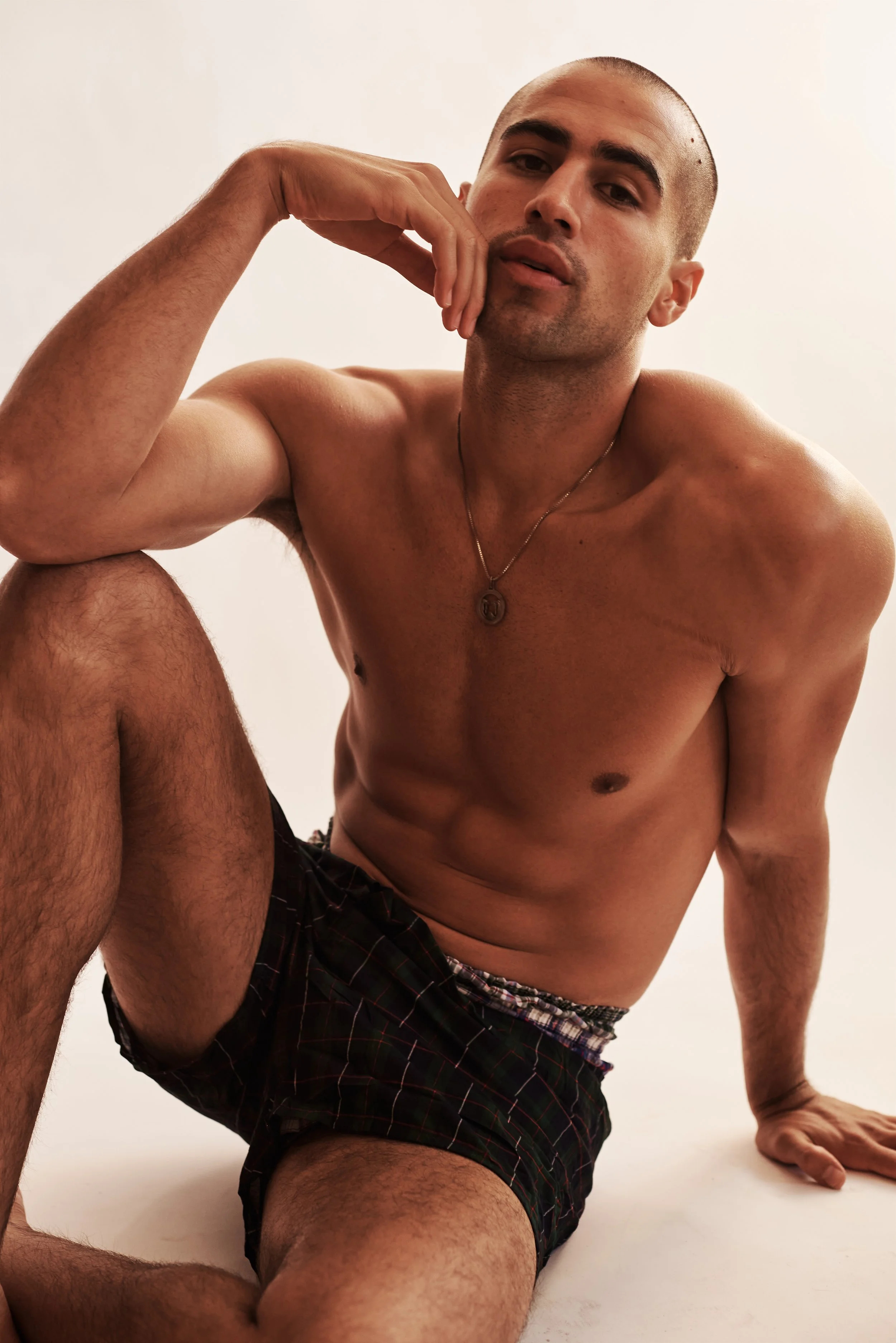 Studio test shoot of a shirtless man seated on the floor in patterned shorts, soft light on a pale backdrop.