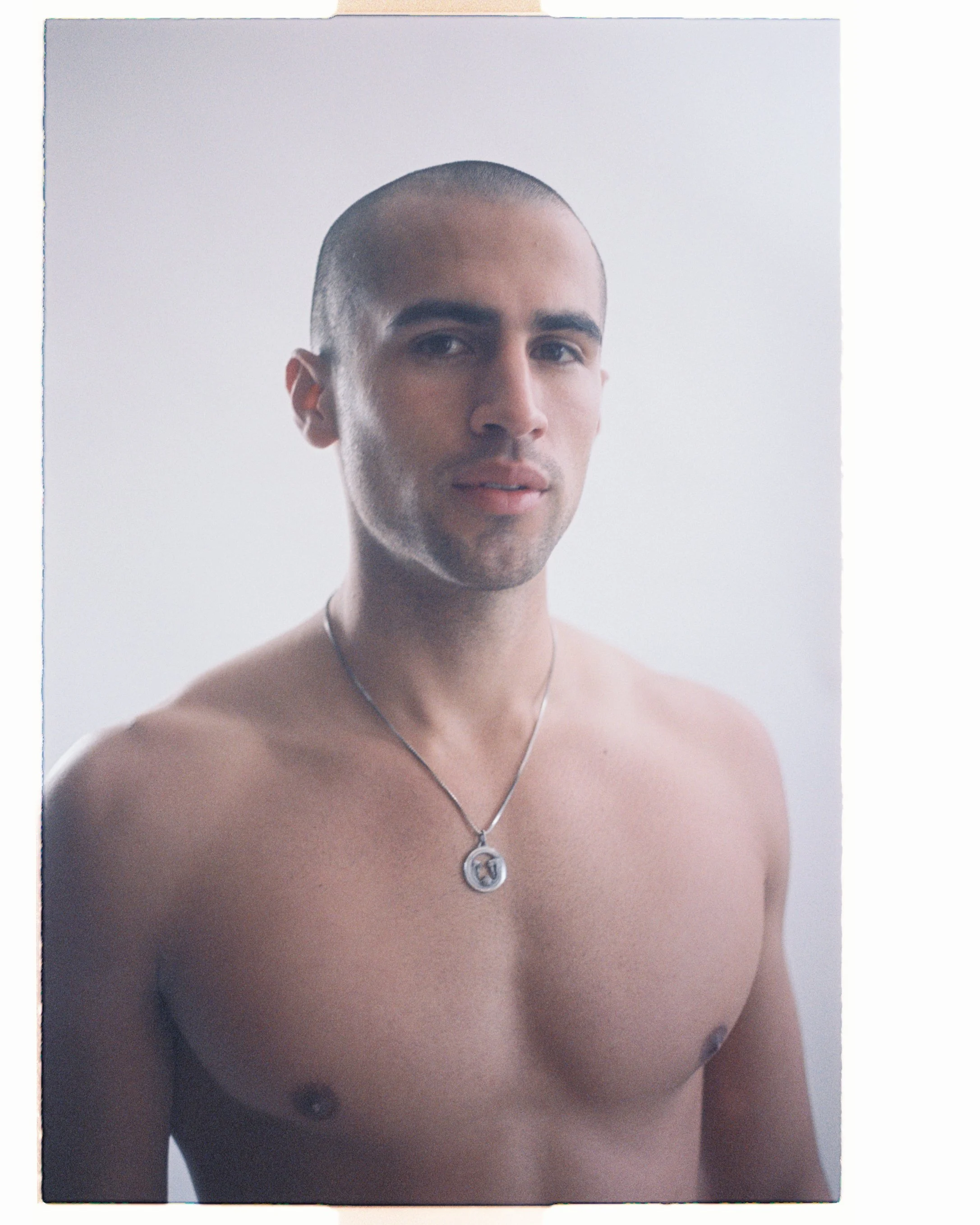 Soft-lit studio test portrait of a shirtless man with a shaved head and a small pendant necklace on a white backdrop.