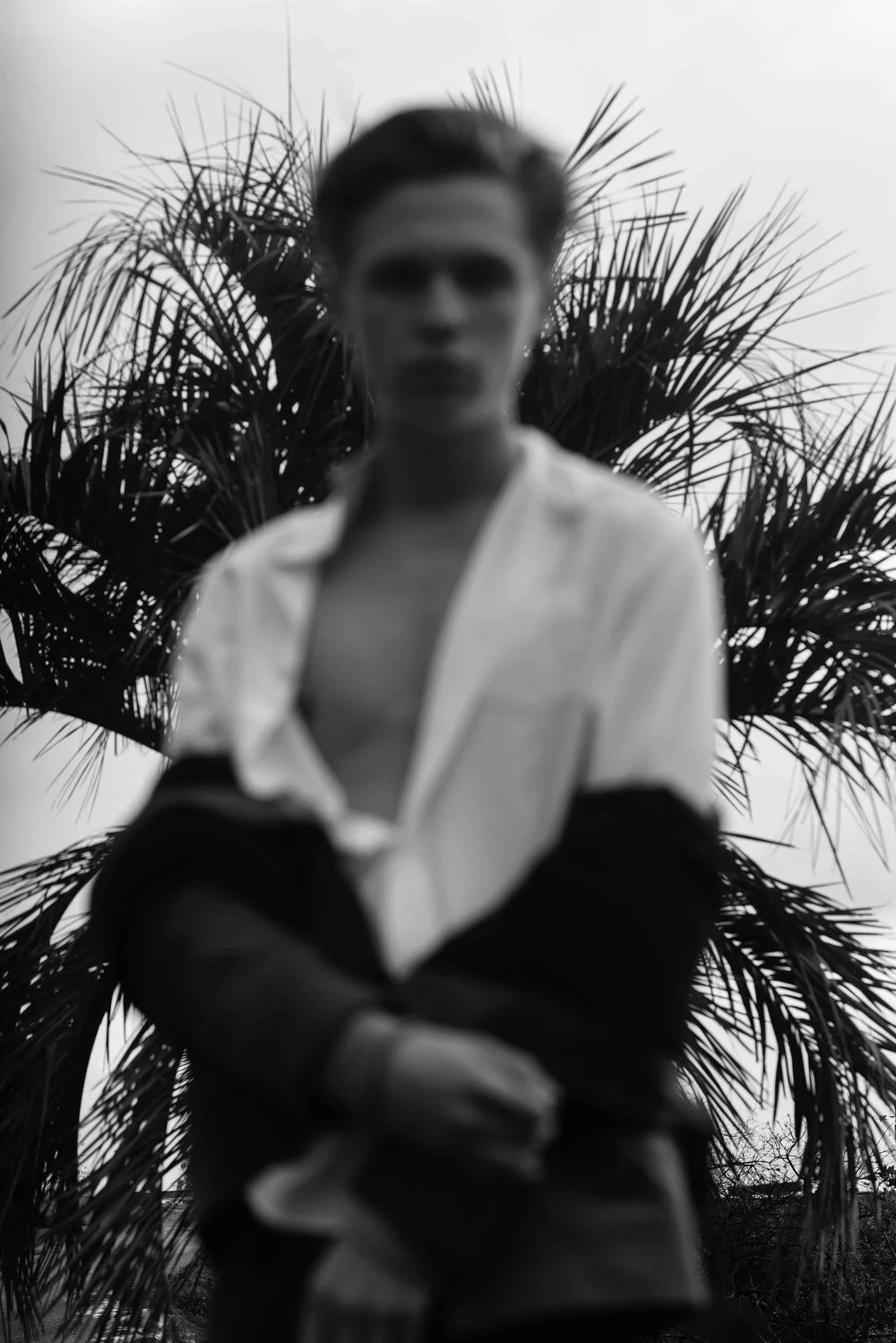 Soft-focus black-and-white test frame of a man in an open white shirt standing in front of palm fronds.