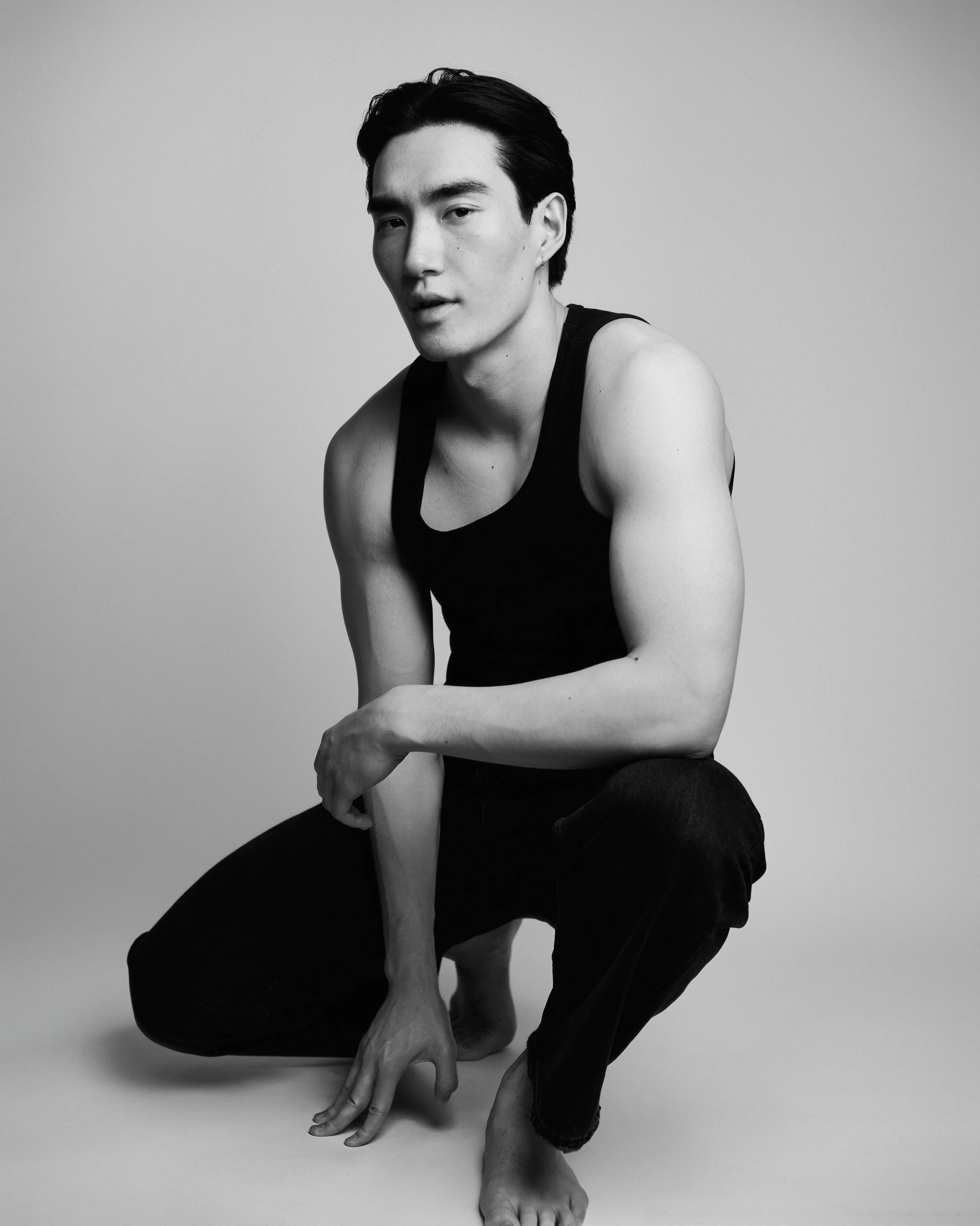 Black-and-white studio test shoot of a man in a black tank top and trousers crouching on a seamless grey backdrop.