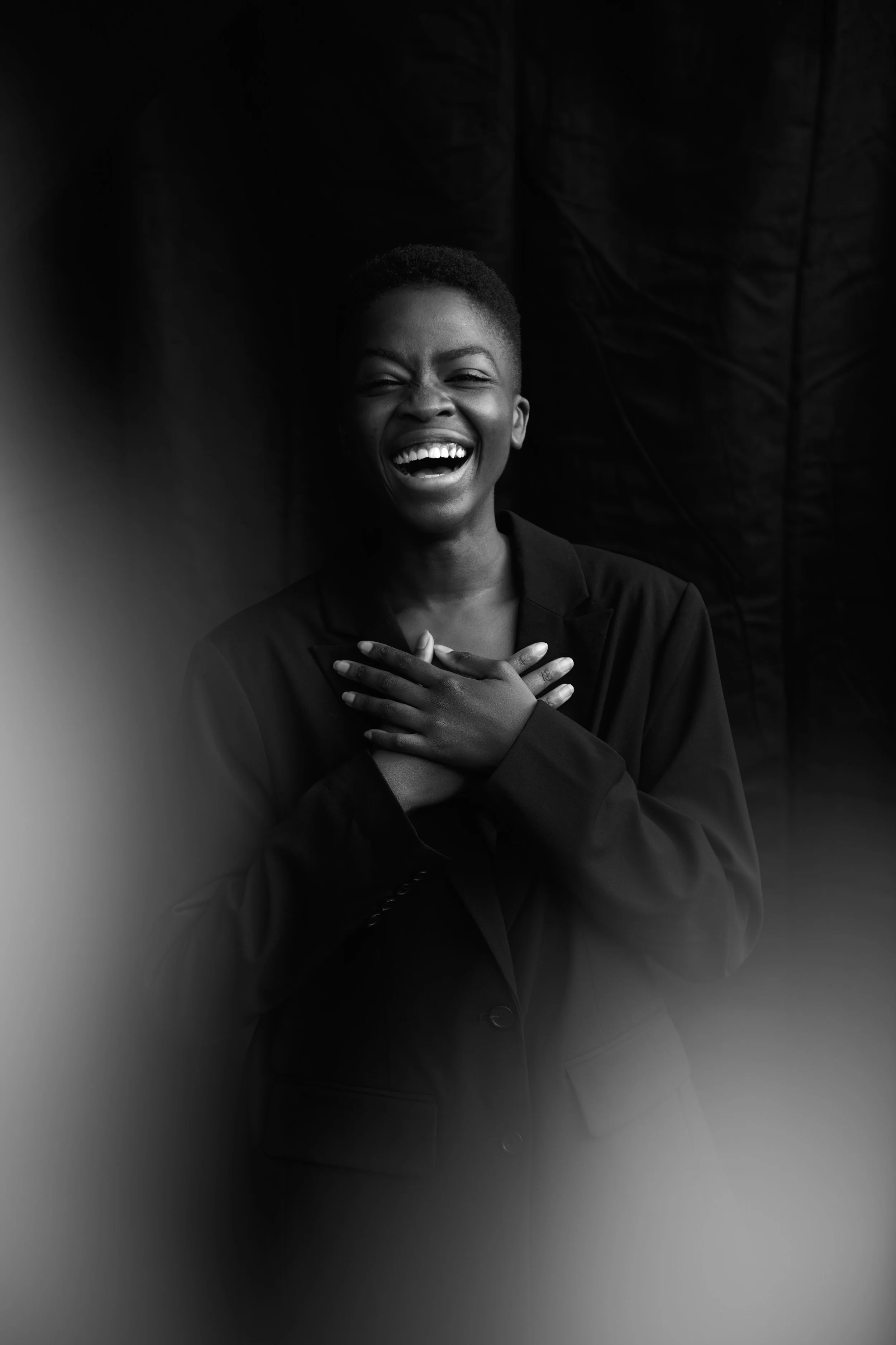 Black-and-white studio test portrait of a laughing woman in a black blazer, hands crossed at her chest, dark backdrop.