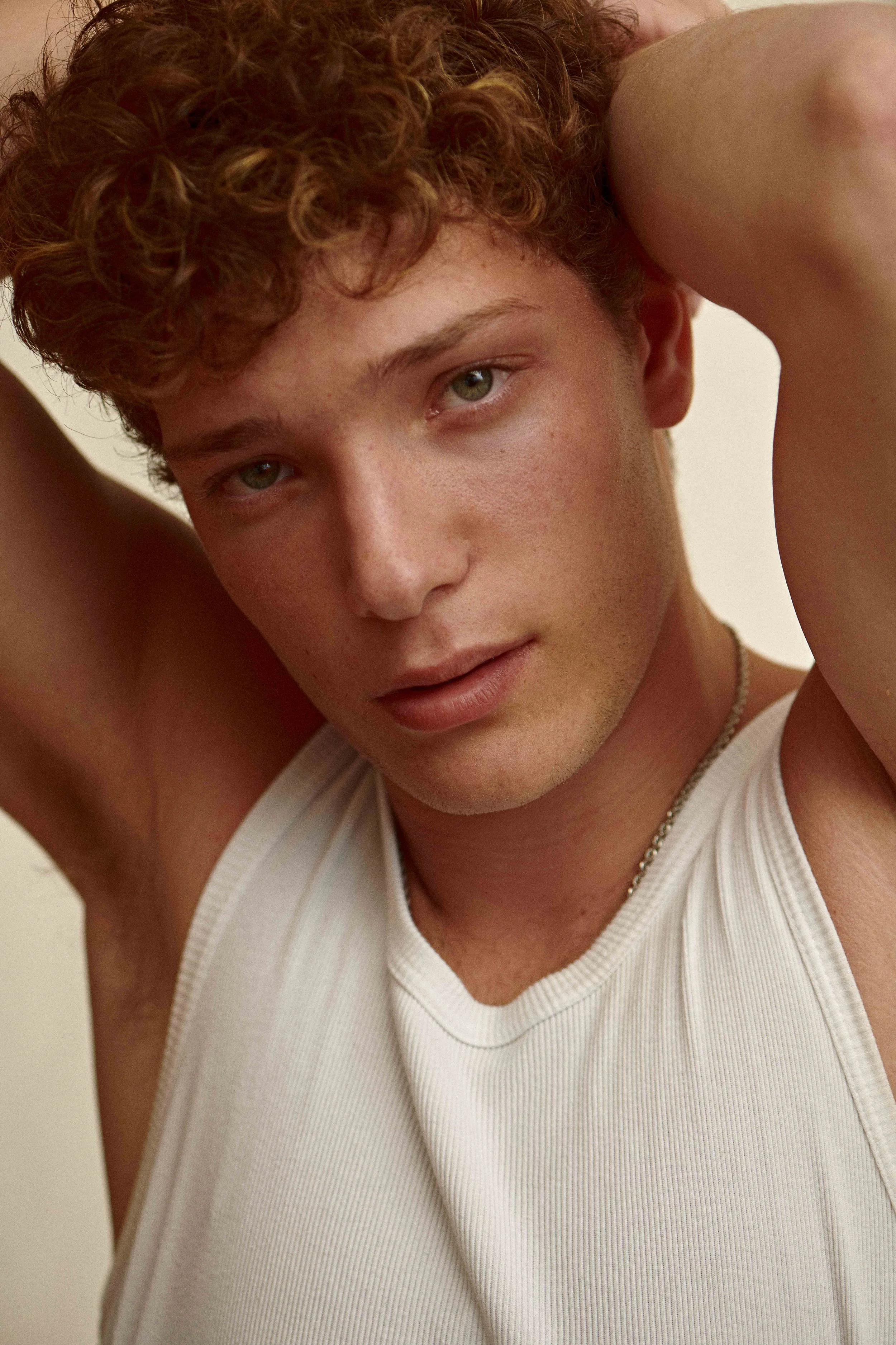 Natural-light beauty test portrait of a young man with red curly hair in a white ribbed tank top, freckles left visible.