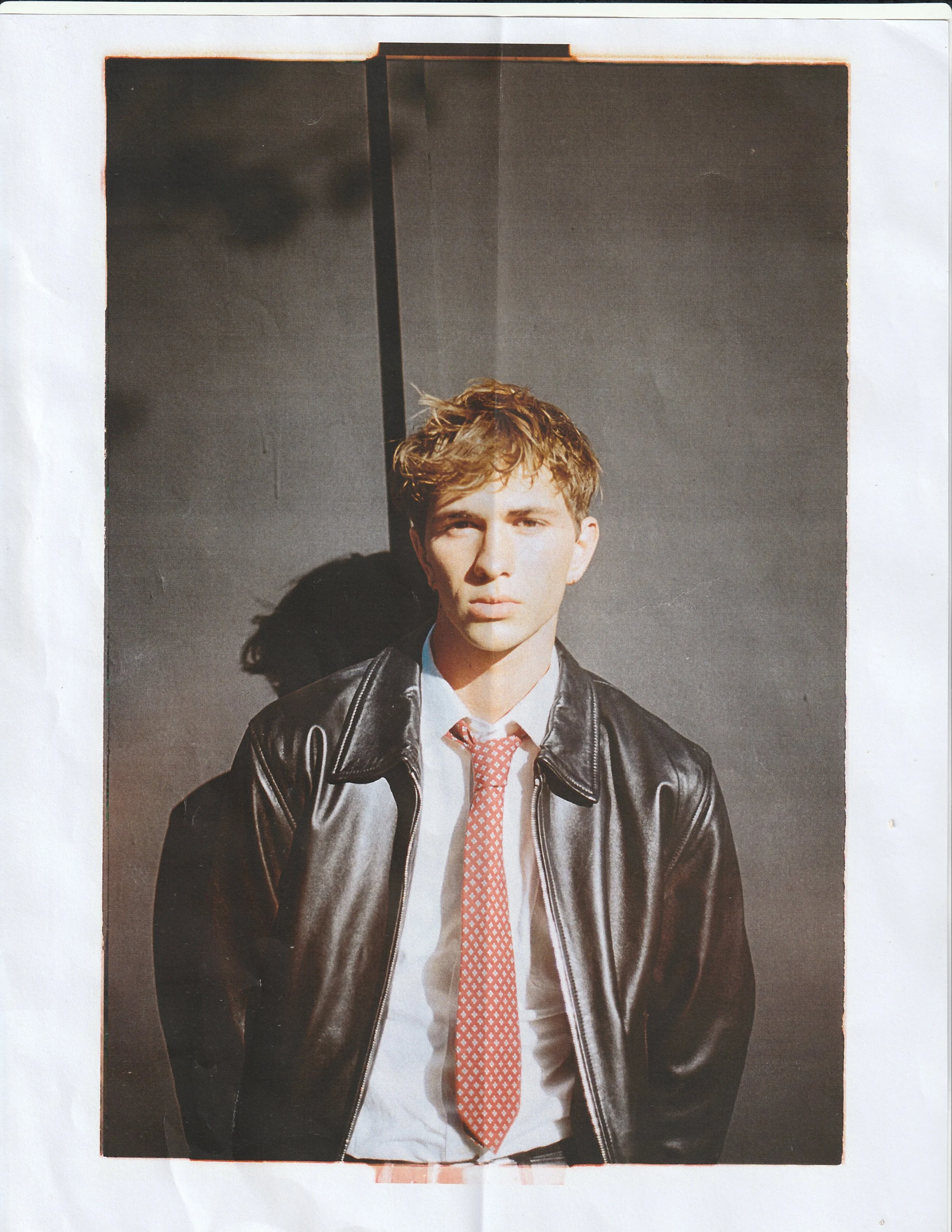 Editorial test portrait of a blond man in a brown leather jacket, white shirt and coral tie, shot against a paper backdrop.