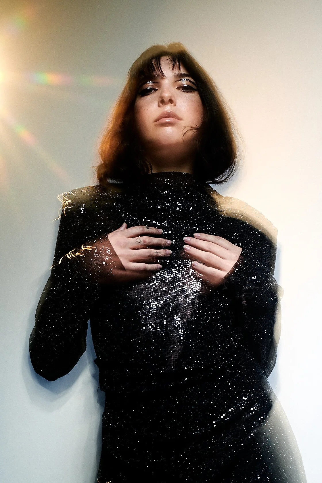 Test shoot of a woman with a dark bob in a black sequinned top, backlit against a pale wall with lens flare.